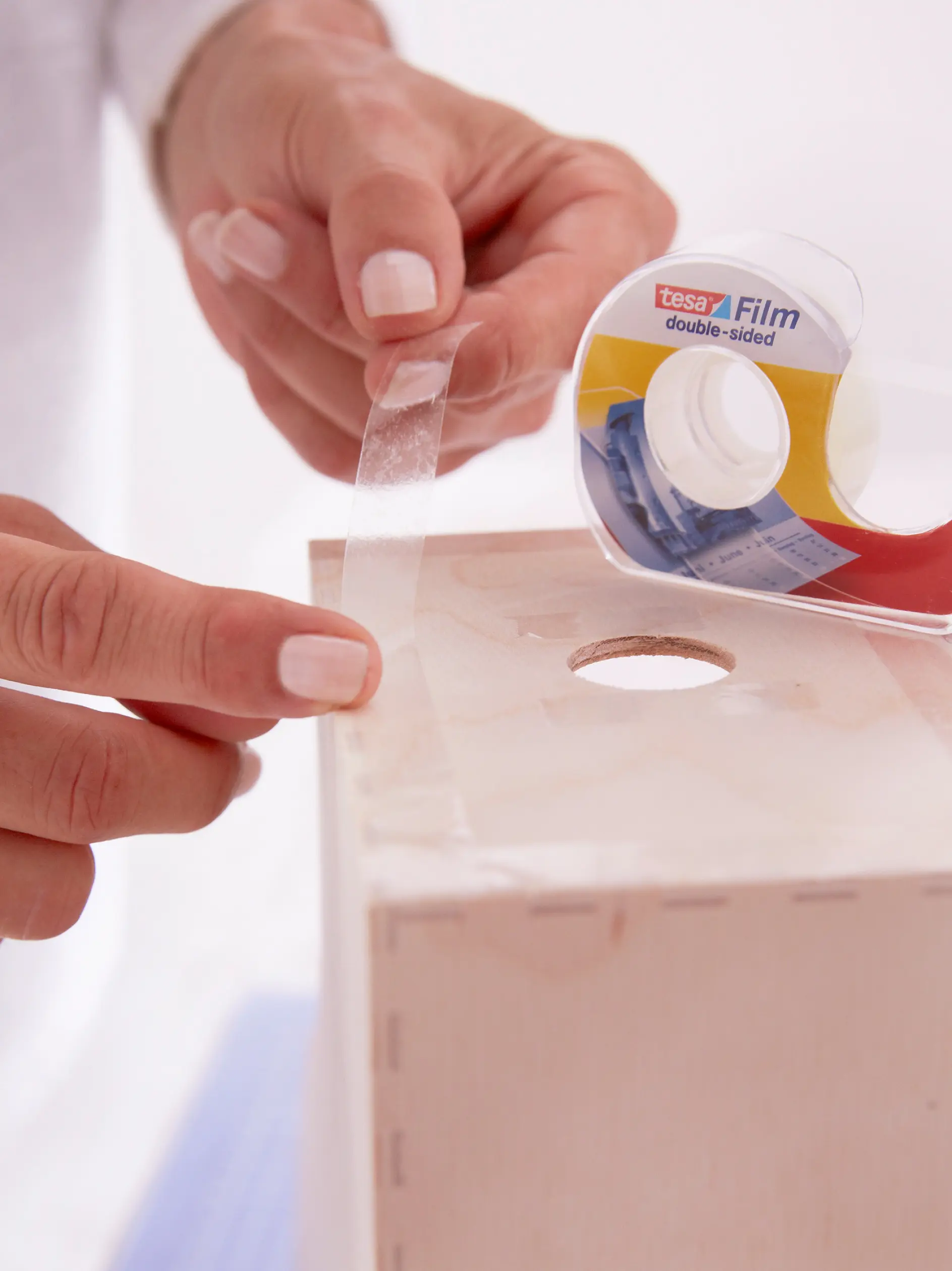 A person applies double-sided tesa tape onto a wooden box using a roll of tesa tape. The person’s hands are visible, and the box has a hole on top. The tesa tape appears translucent, and the persons nails are painted white.