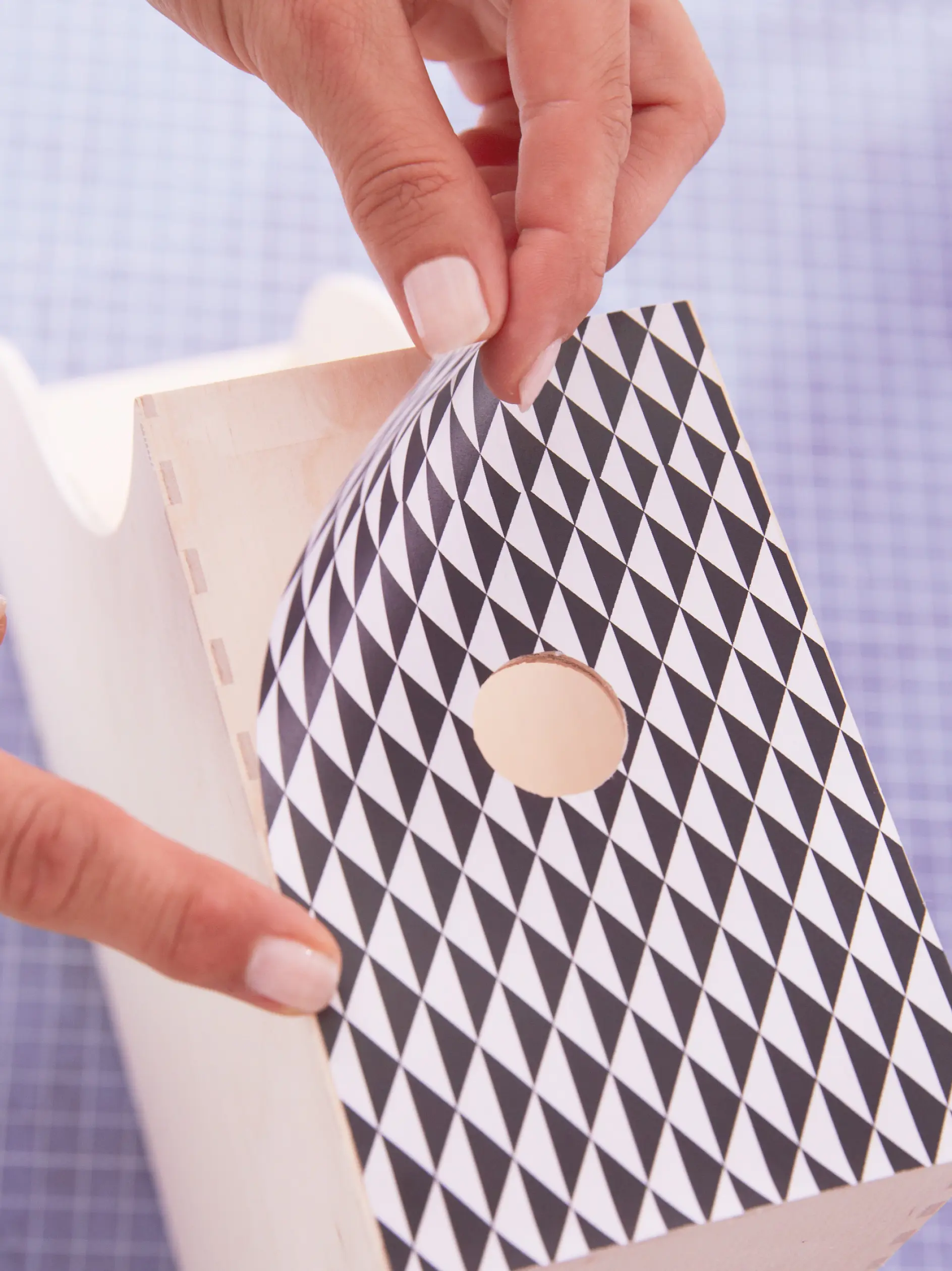 Close-up of hands applying a black and white diamond-patterned adhesive paper to a wooden birdhouse using tesa tape. The birdhouse has a circular opening in the center. The background features a blue grid pattern.
