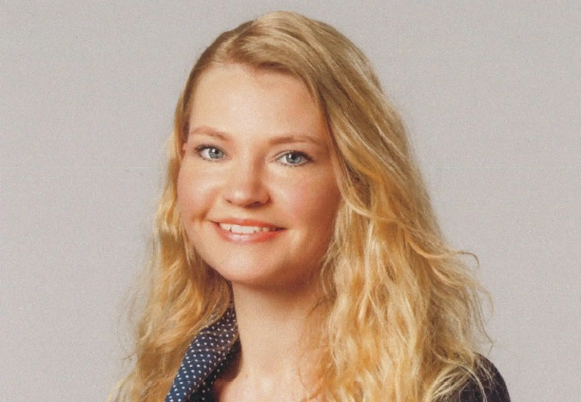 A person with long blonde hair is smiling at the camera. They are wearing a dark blazer with a polka dot shirt underneath. The background is plain and light-colored, featuring tesa tape prominently adhered to various surfaces in the setting.