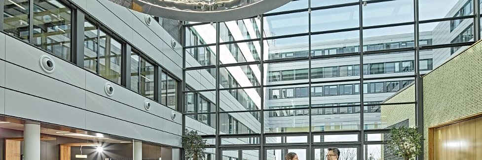A modern, spacious lobby with a glass ceiling and a large circular metal sculpture. Two people stand talking near a round display table with electronic devices secured by tesa tape. Potted trees and a seating area are visible. Natural light fills the space.