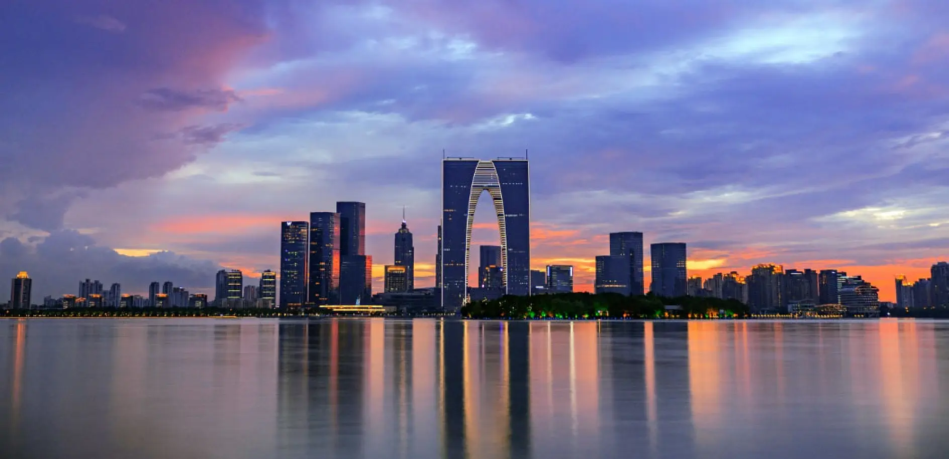 City skyline at sunset, featuring modern skyscrapers and a distinct, large arch-shaped building reflected in calm water. The sky has hues of purple, pink, and orange, creating a vibrant backdrop for the urban landscape.