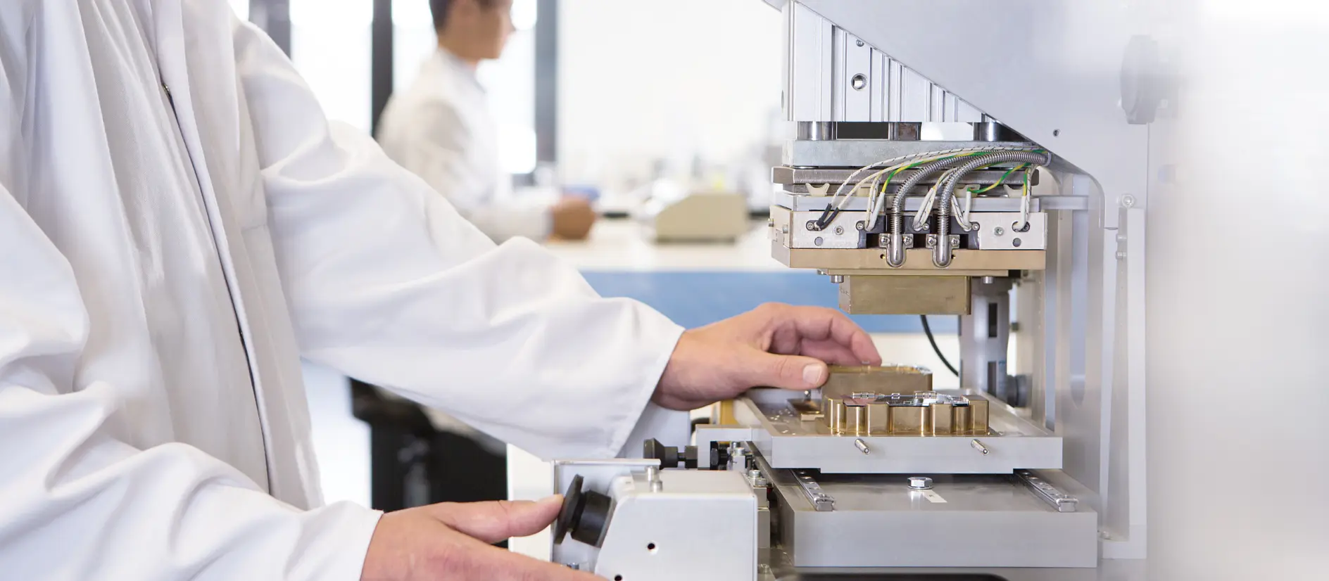 A person in a lab coat operates a scientific instrument with multiple wires and metal components, using tesa tape for secure placement. Another individual works in the background. The setting appears to be a laboratory with a focus on research or testing.