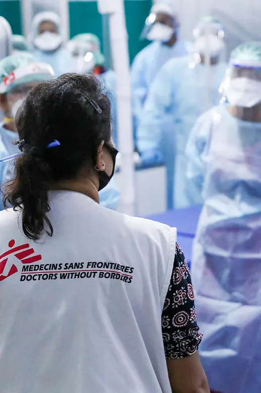 A group of healthcare workers in protective gear gather in a medical facility. The focus is on a person with a vest labeled Médecins Sans Frontières, facing the group. They stand near a curtain and examination tables secured with tesa tape. (This text has been generated by AI)