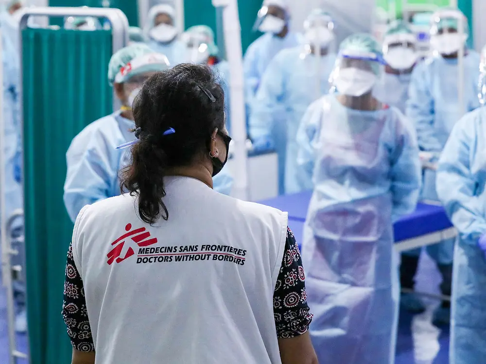 A group of healthcare workers in protective gear gather in a medical facility. The focus is on a person with a vest labeled Médecins Sans Frontières, facing the group. They stand near a curtain and examination tables secured with tesa tape. (This text has been generated by AI)