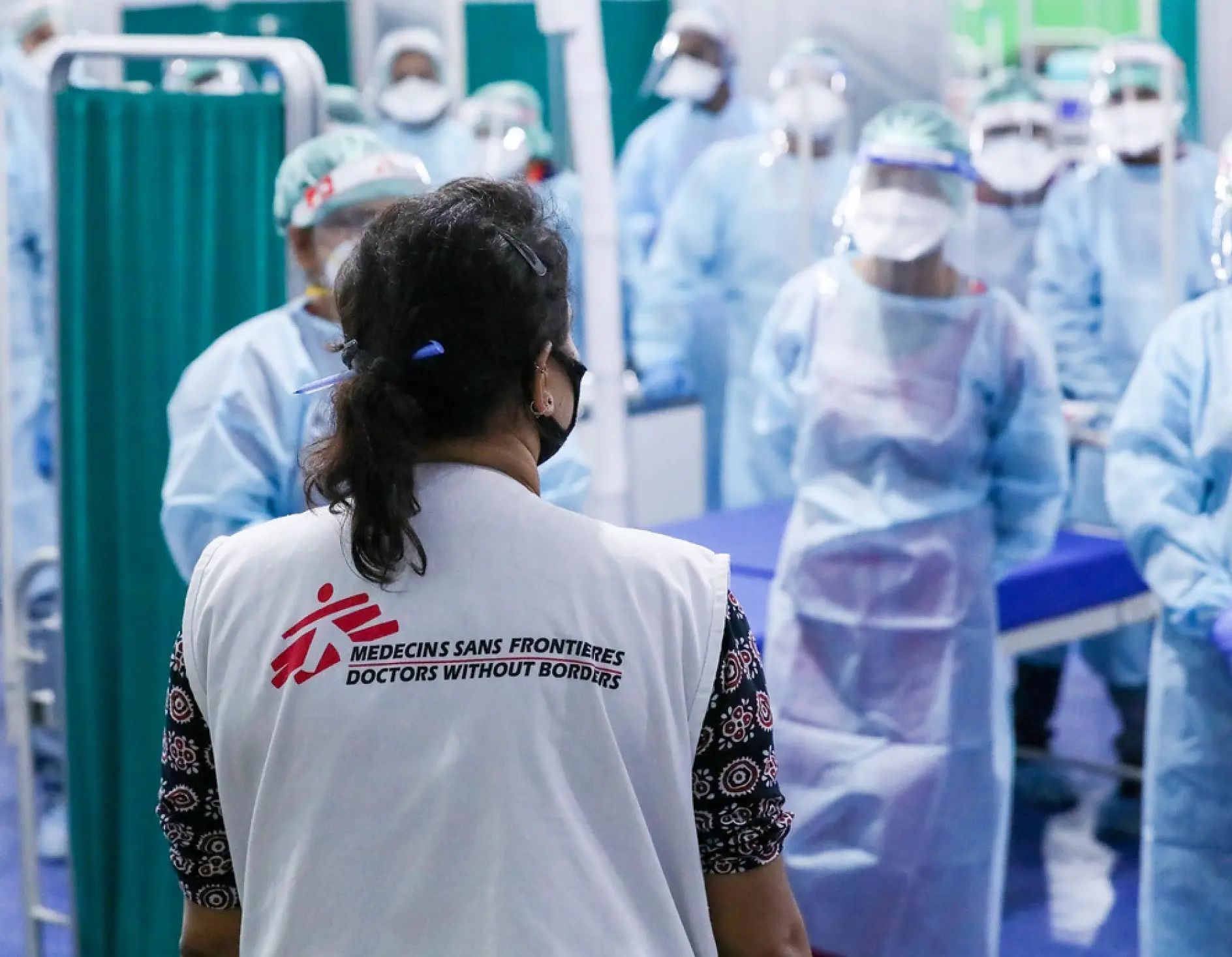 A group of healthcare workers in protective gear gather in a medical facility. The focus is on a person with a vest labeled Médecins Sans Frontières, facing the group. They stand near a curtain and examination tables secured with tesa tape.