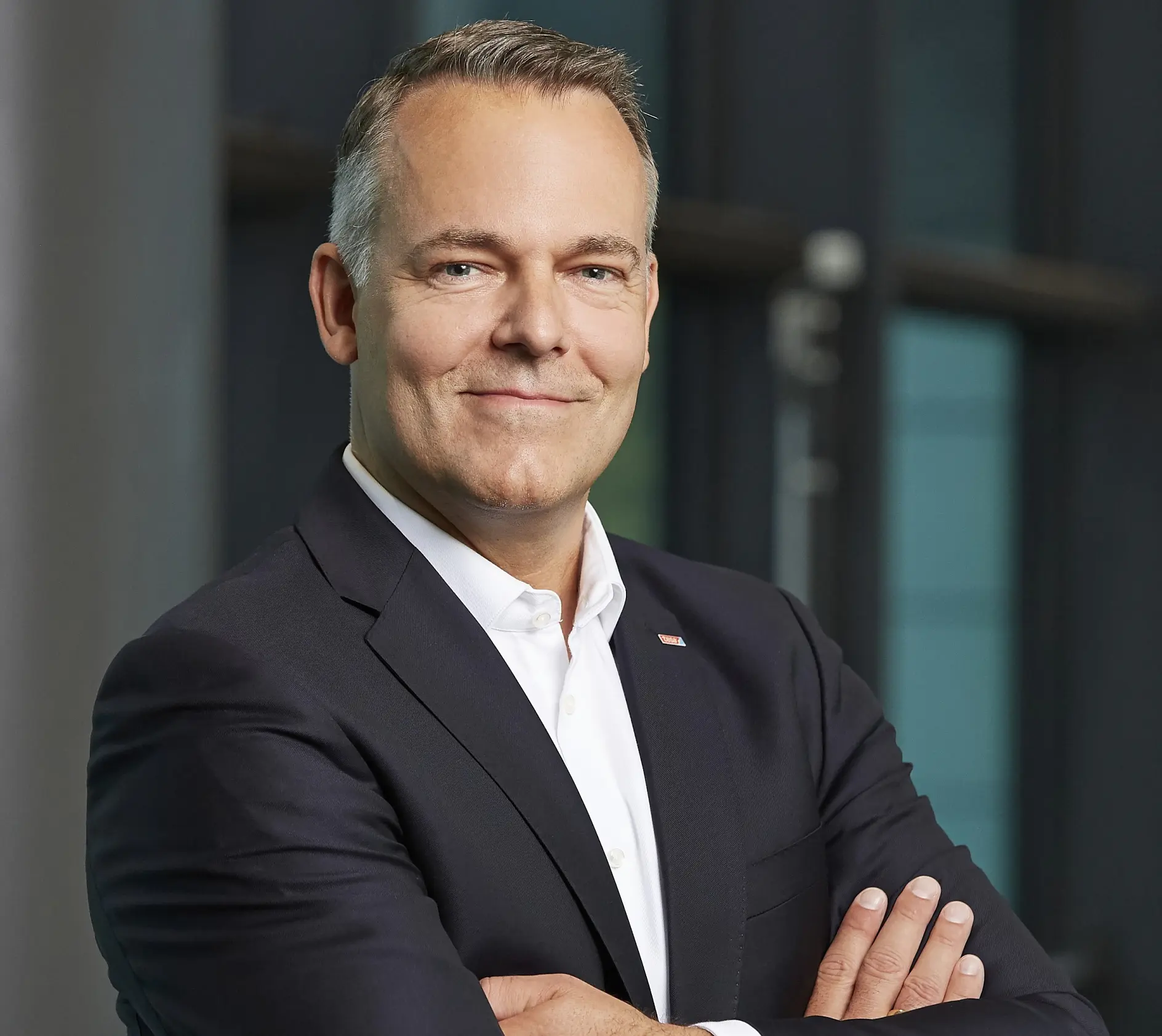 In a dark suit and white shirt, the man smiles with his arms crossed, standing indoors against a blurred background. His short hair is neatly styled, and he has a confident expression.