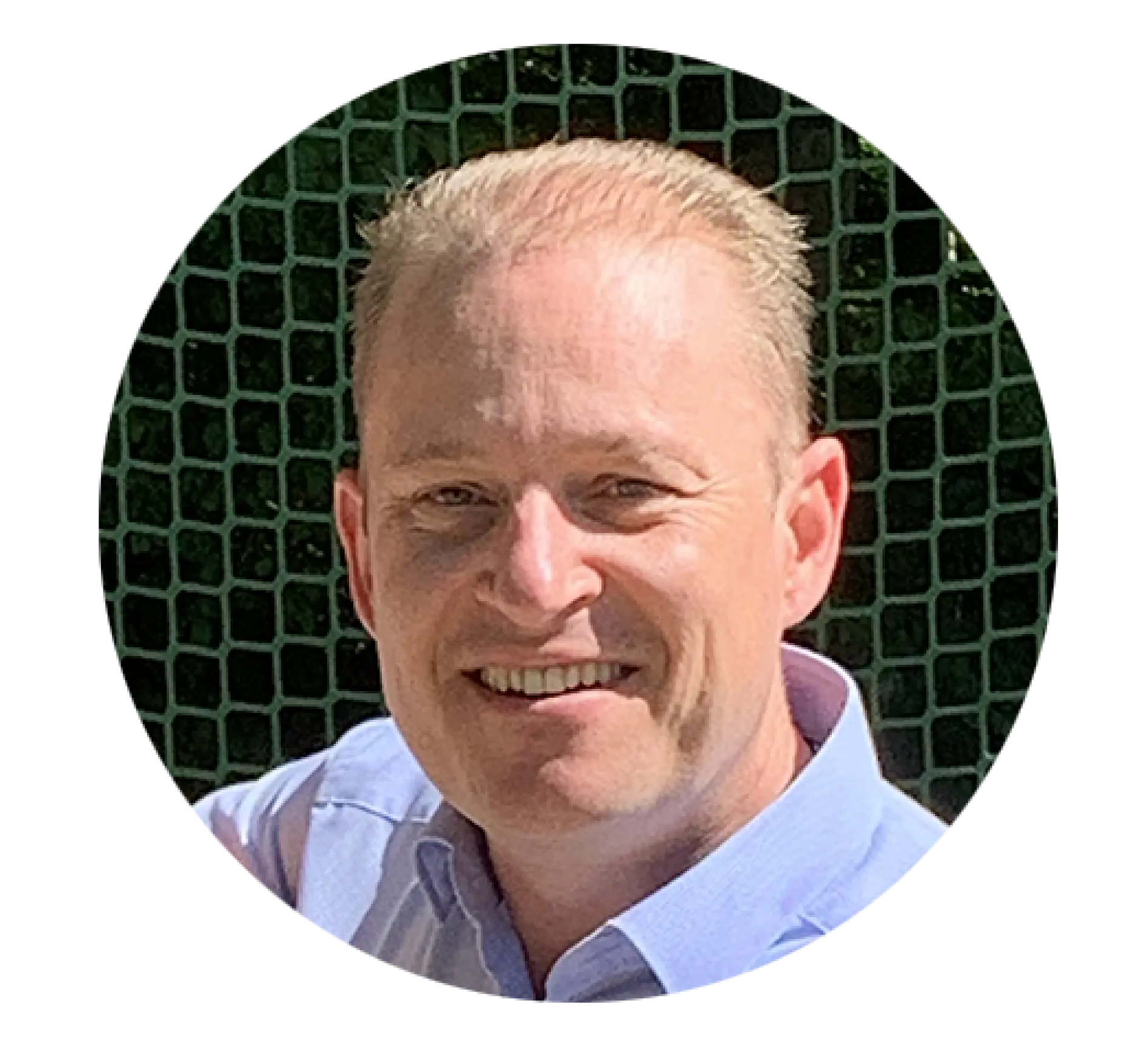 A man with short blonde hair is smiling. He is wearing a light blue collared shirt and is standing in front of a green netted background. The photo is taken in bright daylight.