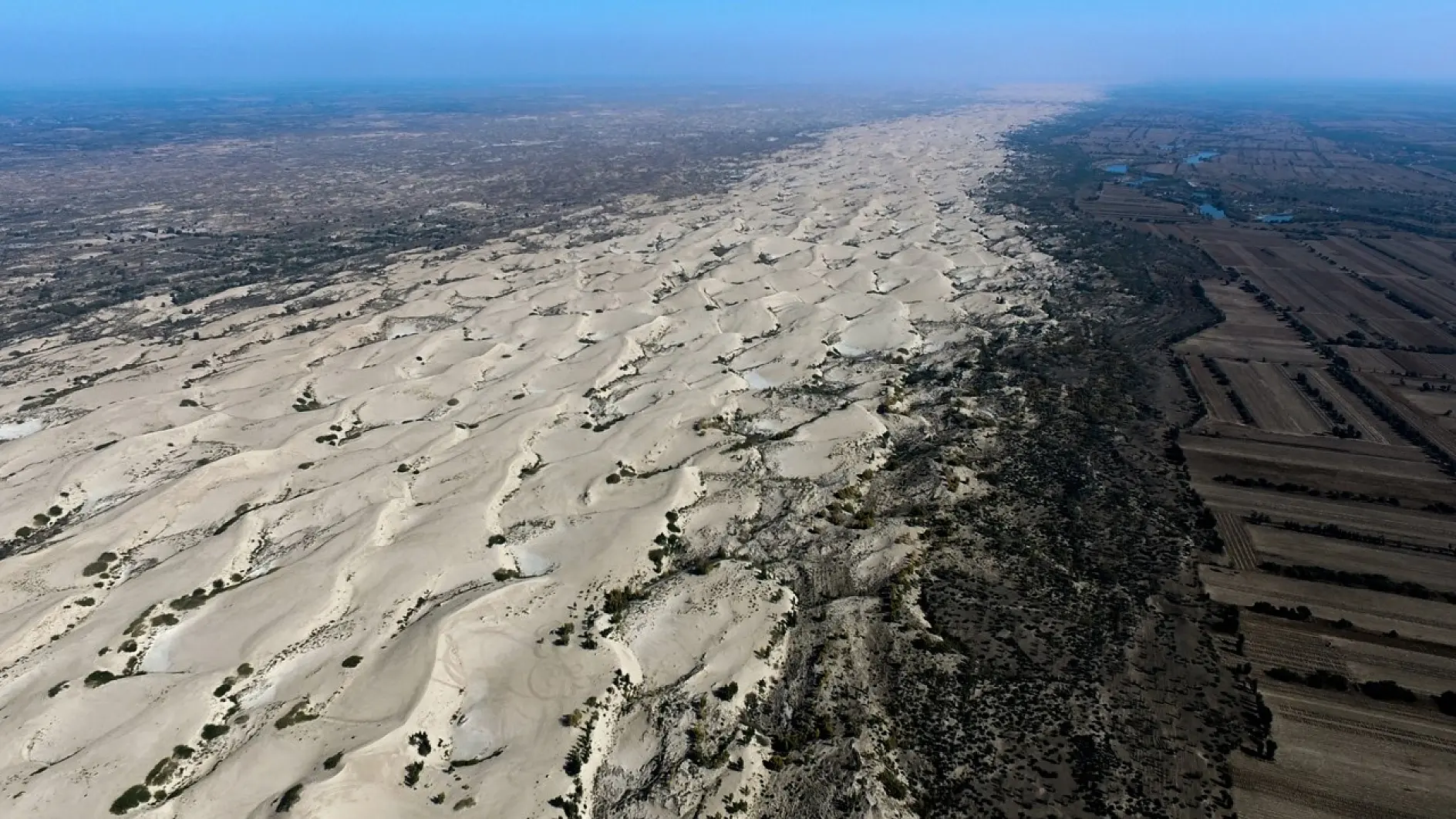 Aerial view of a vast desert landscape with sand dunes stretching across the image. On one side, the dunes transition into a flatter, darker terrain with sparse vegetation. Clear blue sky above.