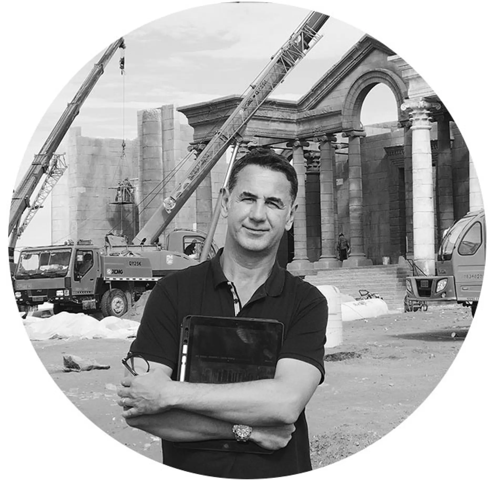 A man stands in front of a construction site with cranes and a partially built classical-style structure. He is holding a tablet and seems to be observing the progress. The image is in black and white and framed in a circular shape.