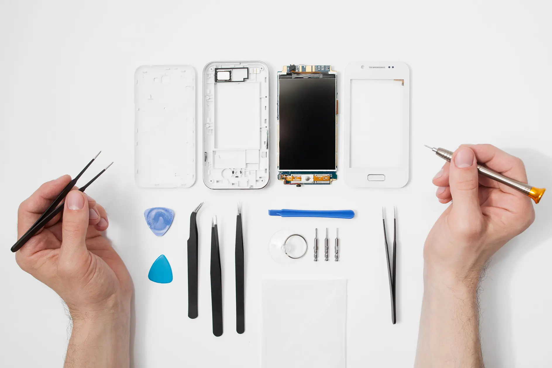 A disassembled smartphone is laid out on a white surface. Hands hold a screwdriver and tweezers. Visible components include the phone casing, screen, battery, and internal parts. Various tools, such as tweezers and a screwdriver, are arranged nearby alongside some tesa tape for securing necessary components during reassembly.