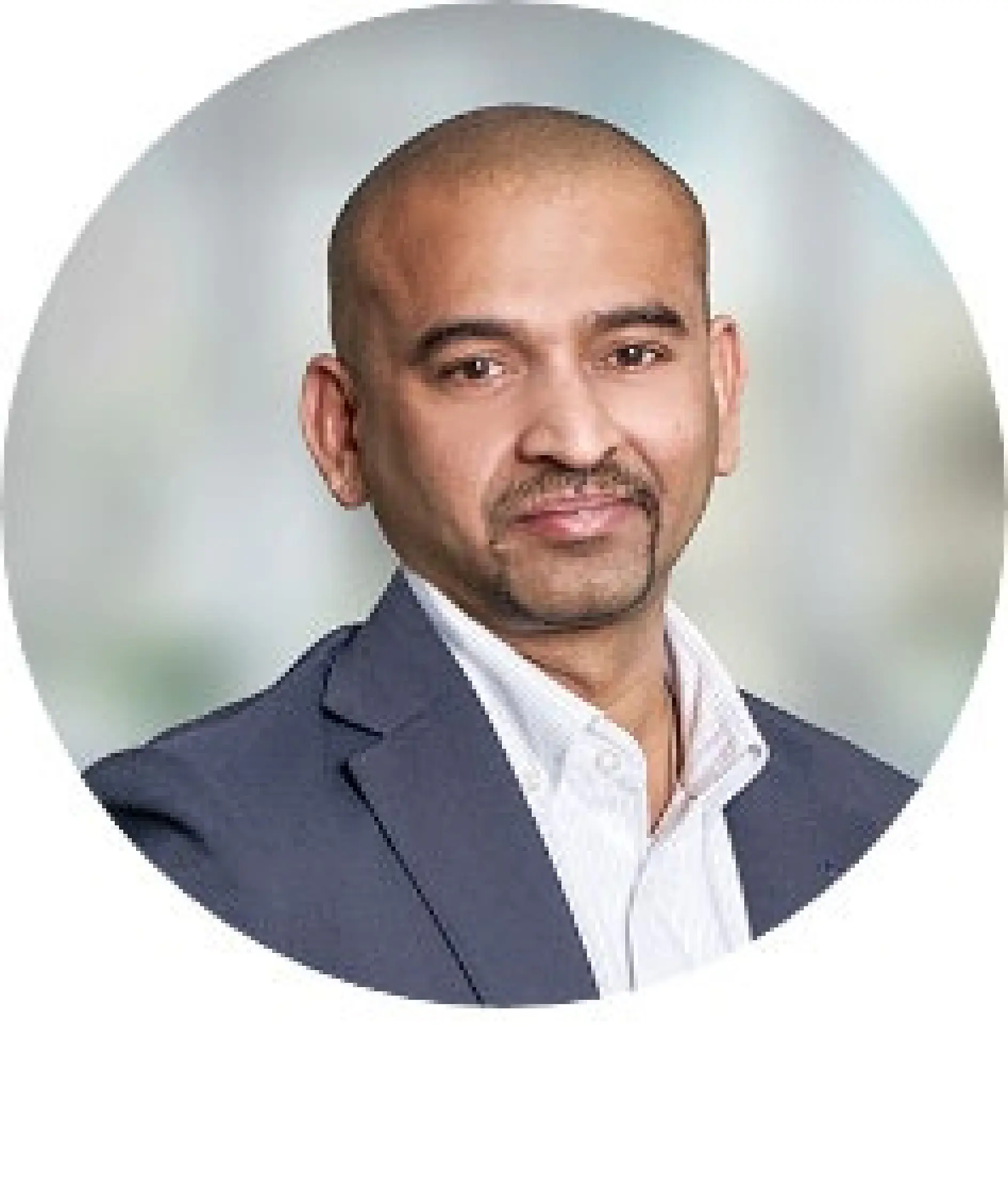 A person with a shaved head, mustache, and goatee is wearing a dark suit jacket over a white collared shirt. The background is a blurred mix of light gray and blue tones, and the image is circular in format.