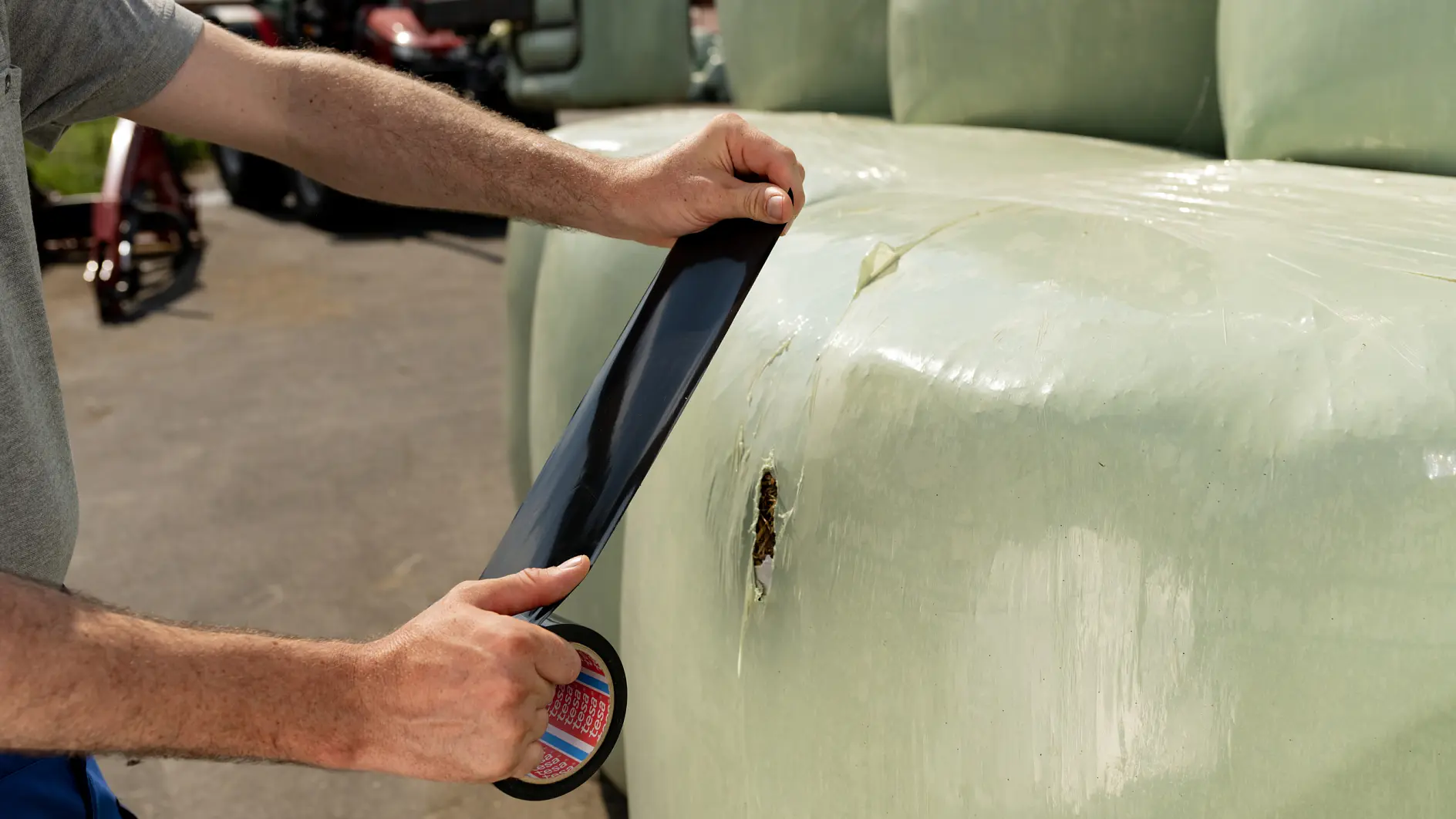 A person repairs a damaged spot on a large wrapped bale of hay with black tesa tape. The bale is covered with a light green plastic. Farm machinery can be seen in the background.