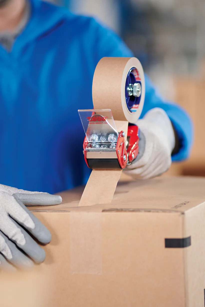 A person wearing a blue jacket and gray gloves is sealing a cardboard box with brown packing tesa tape using a red tesa tape dispenser. Other cardboard boxes are visible in the background. (This text has been generated by AI)