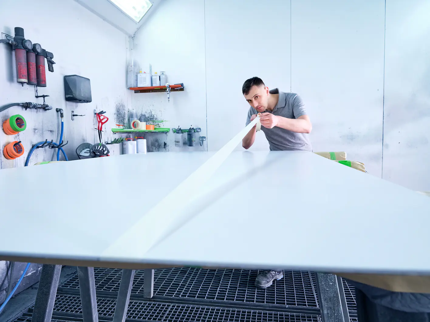 A man in a gray t-shirt is working in a brightly lit workshop. He is peeling white tesa tape from a large, flat surface on a metal table. Various tools and equipment are visible in the background. (This text has been generated by AI)