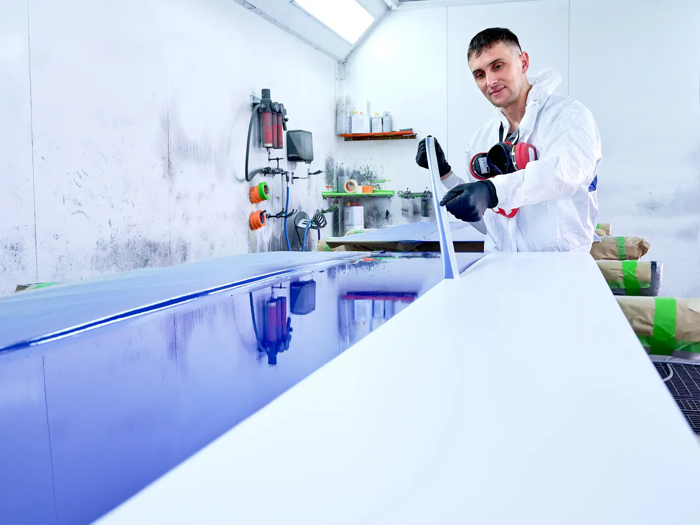 A person in protective clothing spray paints a large panel in a workshop. The room is equipped with various tools and paint supplies, including tesa tape. The panel is half-painted in blue, while the other half remains white. (This text has been generated by AI)