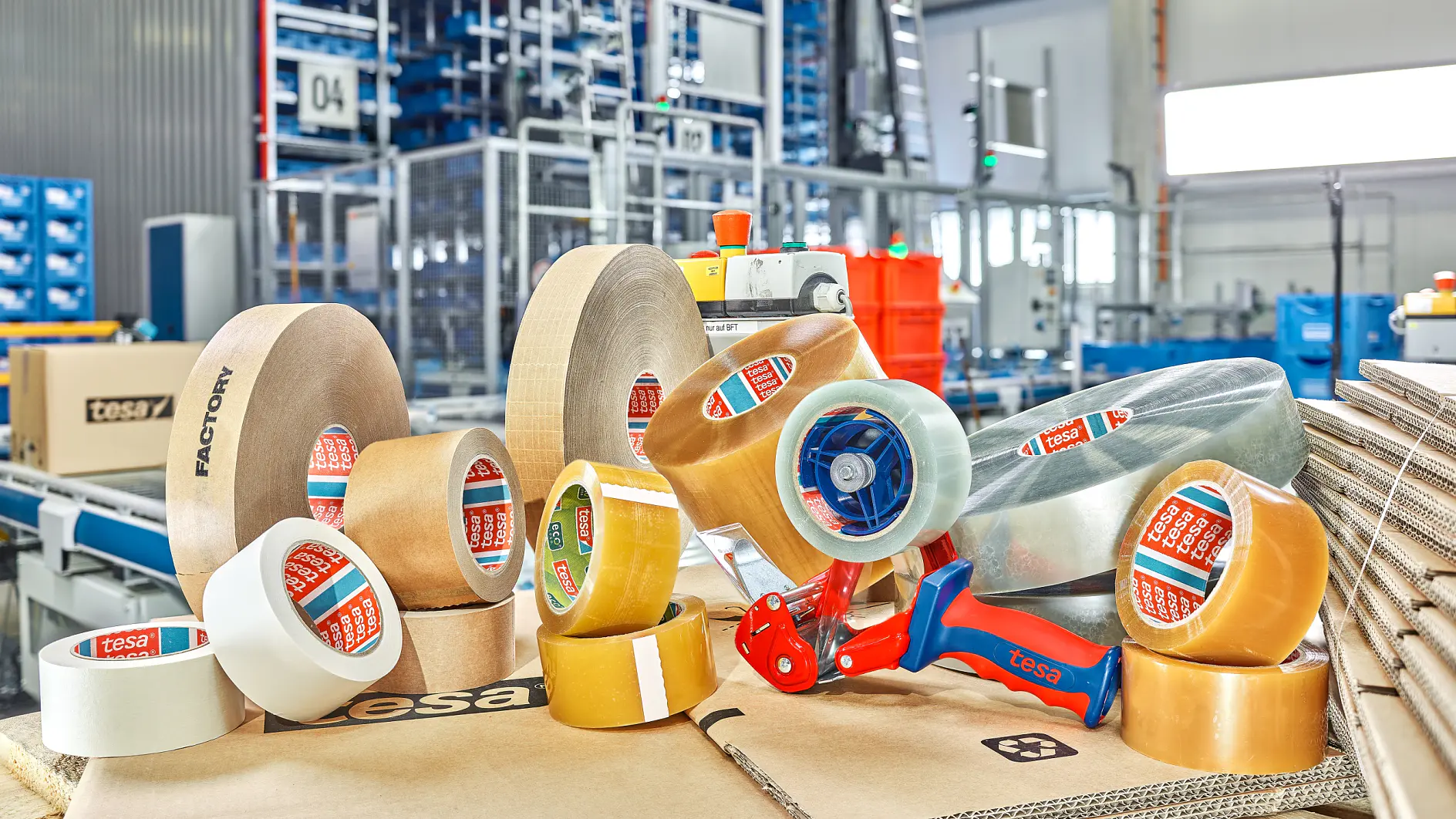 A variety of packing tesa tapes, including clear and brown rolls, are displayed on top of stacked cardboard boxes in an industrial setting. A blue tesa tape dispenser is also present. Shelving and equipment are visible in the background. (This text has been generated by AI)