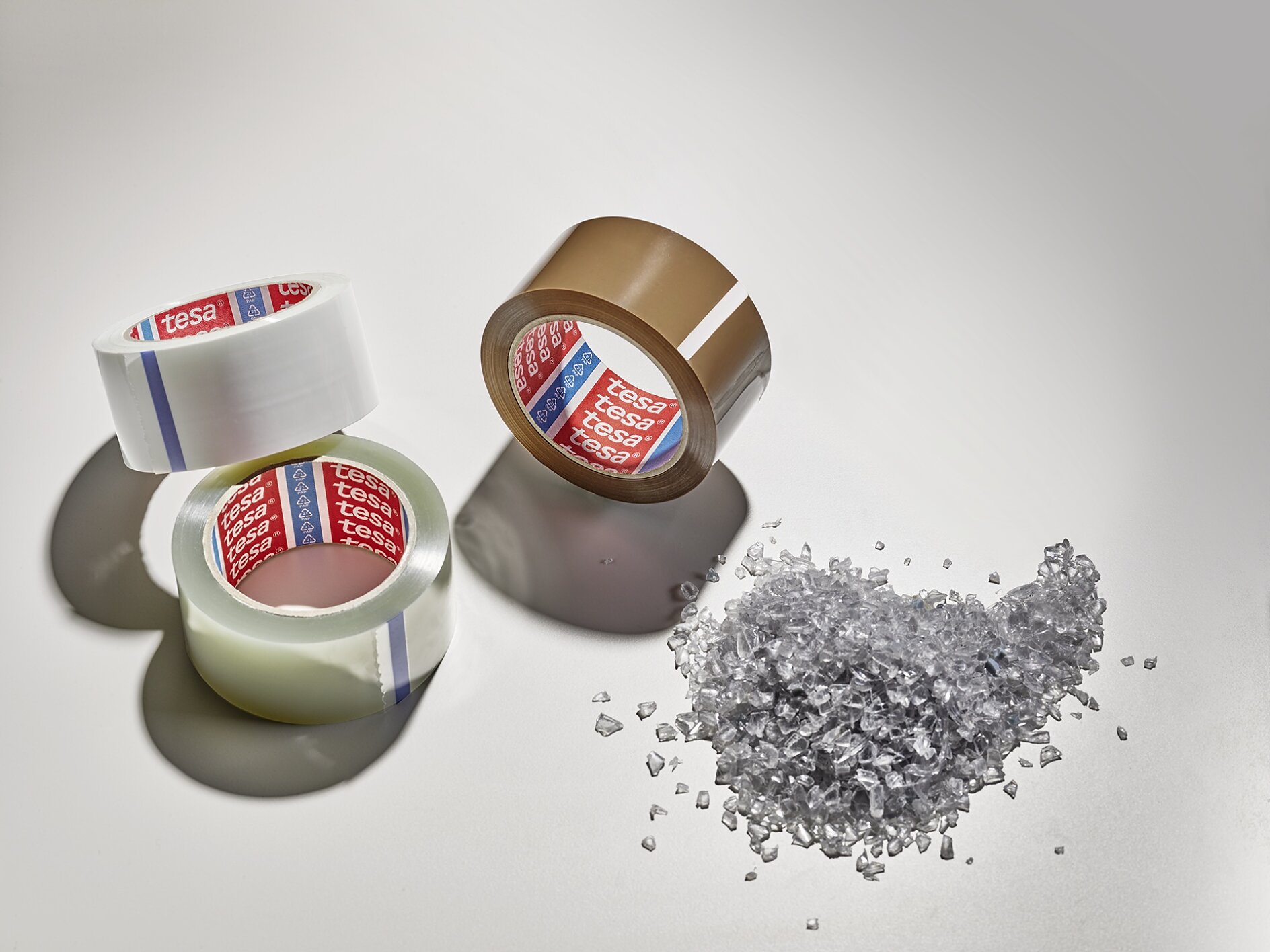 Three rolls of packing tesa tape, two clear and one brown, are arranged on a light surface. Beside them is a small pile of clear plastic granules. Shadows extend from the objects. (This text has been generated by AI)