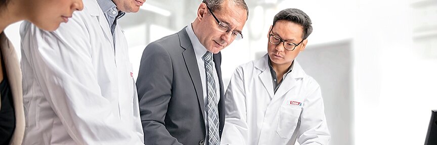 Four people are gathered around a table in a bright office. Two are wearing lab coats, one is in a suit, and one is partially visible. They are focused on documents laid out on the table, which include diagrams labeled with tesa tape. A laptop displaying pertinent data is open nearby. (This text has been generated by AI)