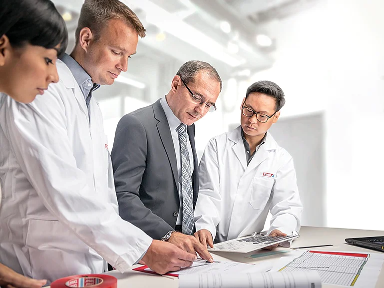 Four people are gathered around a table in a bright office. Two are wearing lab coats, one is in a suit, and one is partially visible. They are focused on documents laid out on the table, which include diagrams labeled with tesa tape. A laptop displaying pertinent data is open nearby. (This text has been generated by AI)