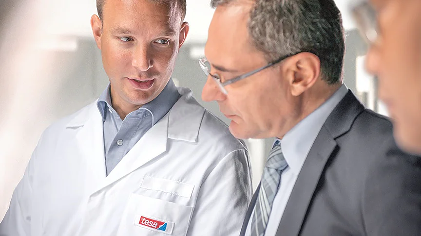 A man in a white lab coat with a tesa tape label talks to another man in a suit and glasses. They appear to be in a discussion while looking at something out of the frame. The background is bright and slightly blurred. (This text has been generated by AI)