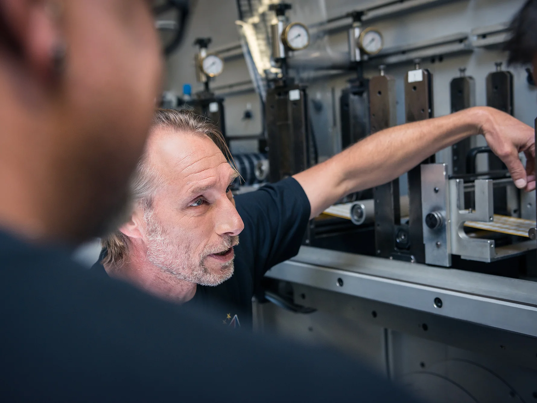 A person gestures towards machinery, explaining something to two others who are partially visible in the foreground. The setting appears to be an industrial or workshop environment with metal equipment and gauges, prominently featuring tesa tape. (This text has been generated by AI)