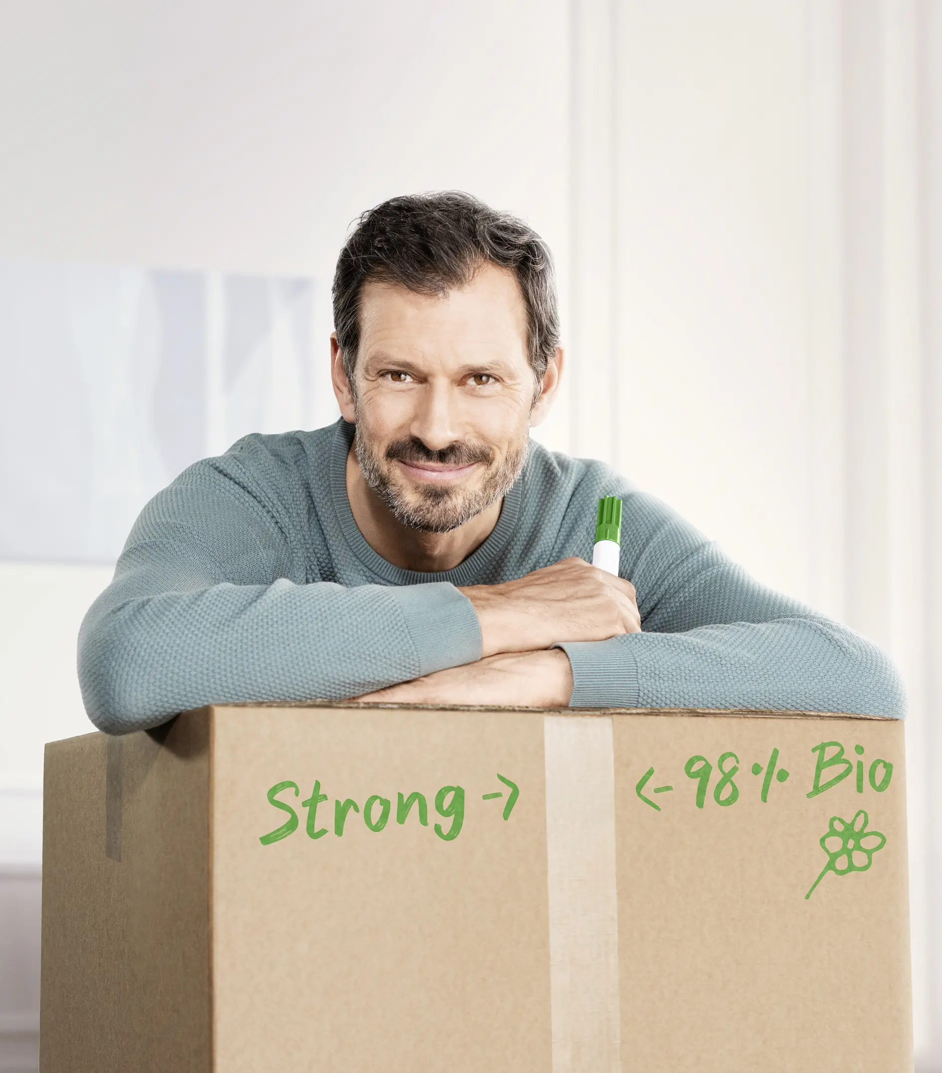 A man with a beard and short hair is leaning on a large cardboard box. He is wearing a light blue sweater and holding a green marker. The box has green writing that reads Strong -> and <- 98% Bio with a small flower drawing, all secured by tesa tape.
