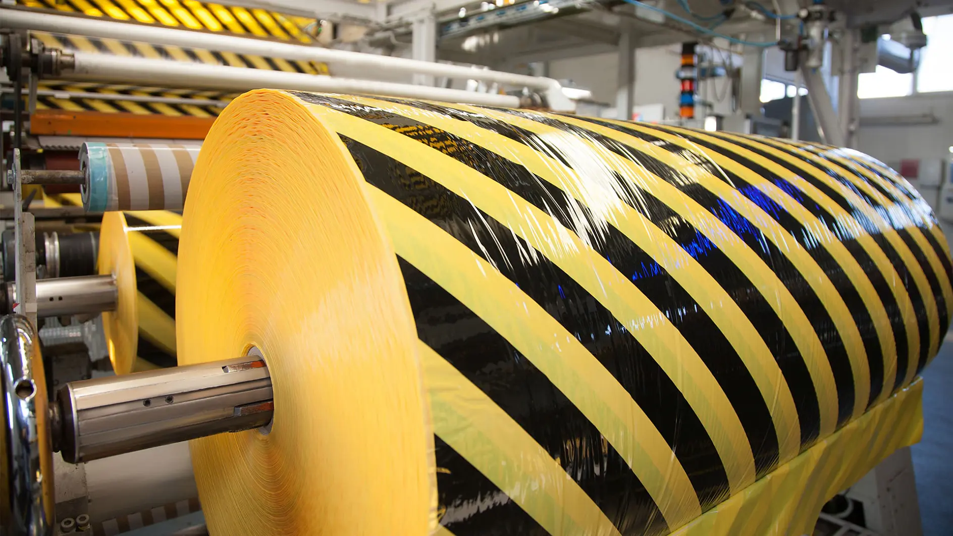A large roll of yellow and black caution tesa tape is mounted on a machine in a factory setting. The tesa tape features diagonal stripes and is ready for processing or cutting. The background shows other rolls and industrial equipment.