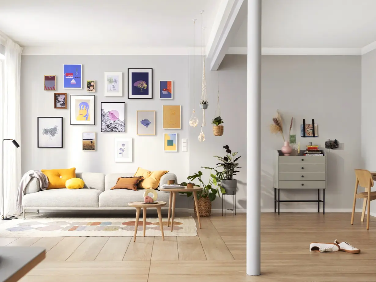 A modern living room with a beige sofa adorned with yellow and gray cushions. Artworks hang on the wall above, secured with tesa tape. A wooden floor extends to an office area with a desk and chair. Houseplants are scattered around, and a soft natural light fills the space.