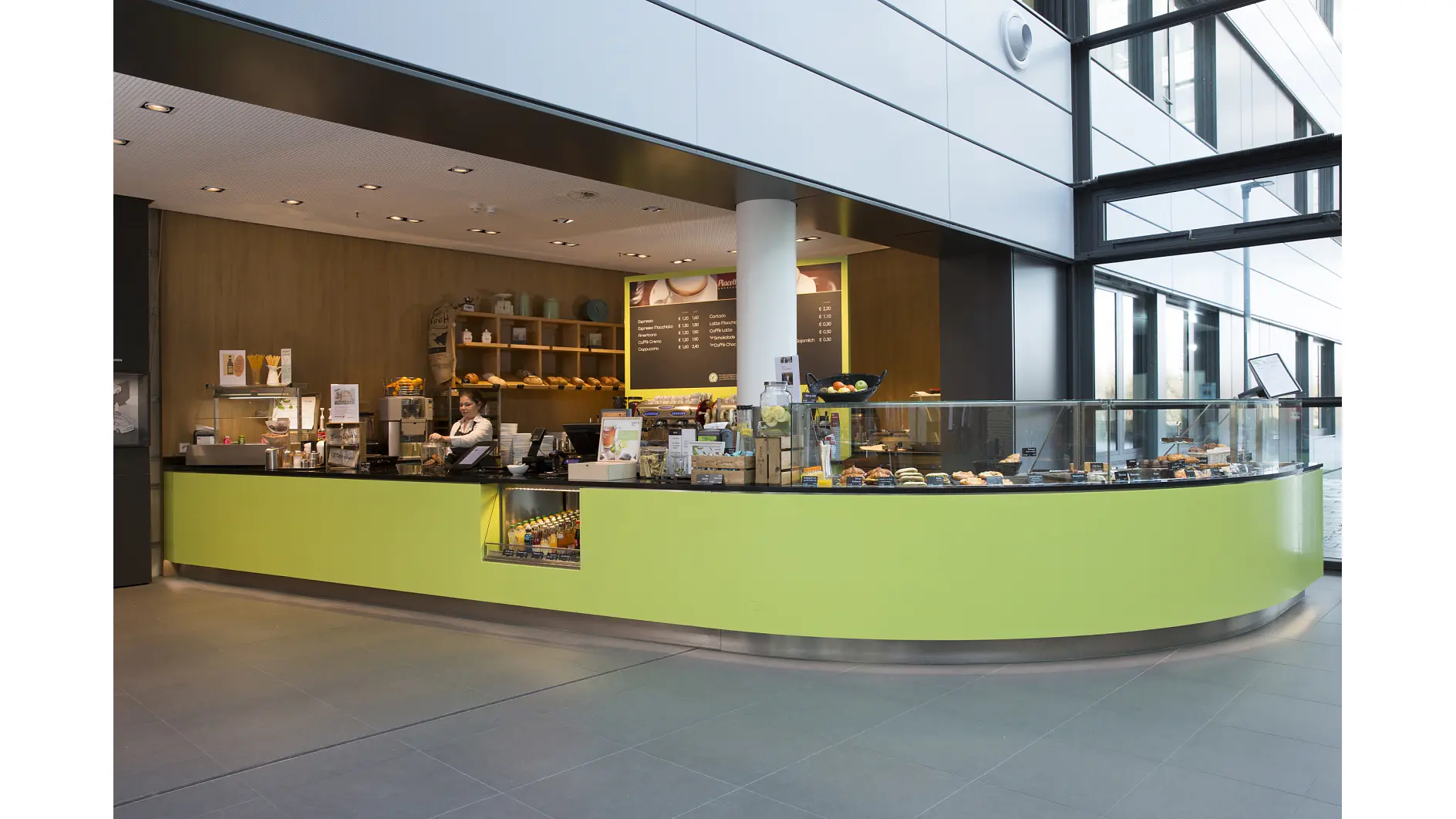 Wide-angle view of a modern café with a long, curved lime-green counter. The menu is displayed on a board behind the counter using tesa tape. Various pastries and drinks are visible. A person stands behind the counter, ready to serve. Large windows add natural light. (This text has been generated by AI)
