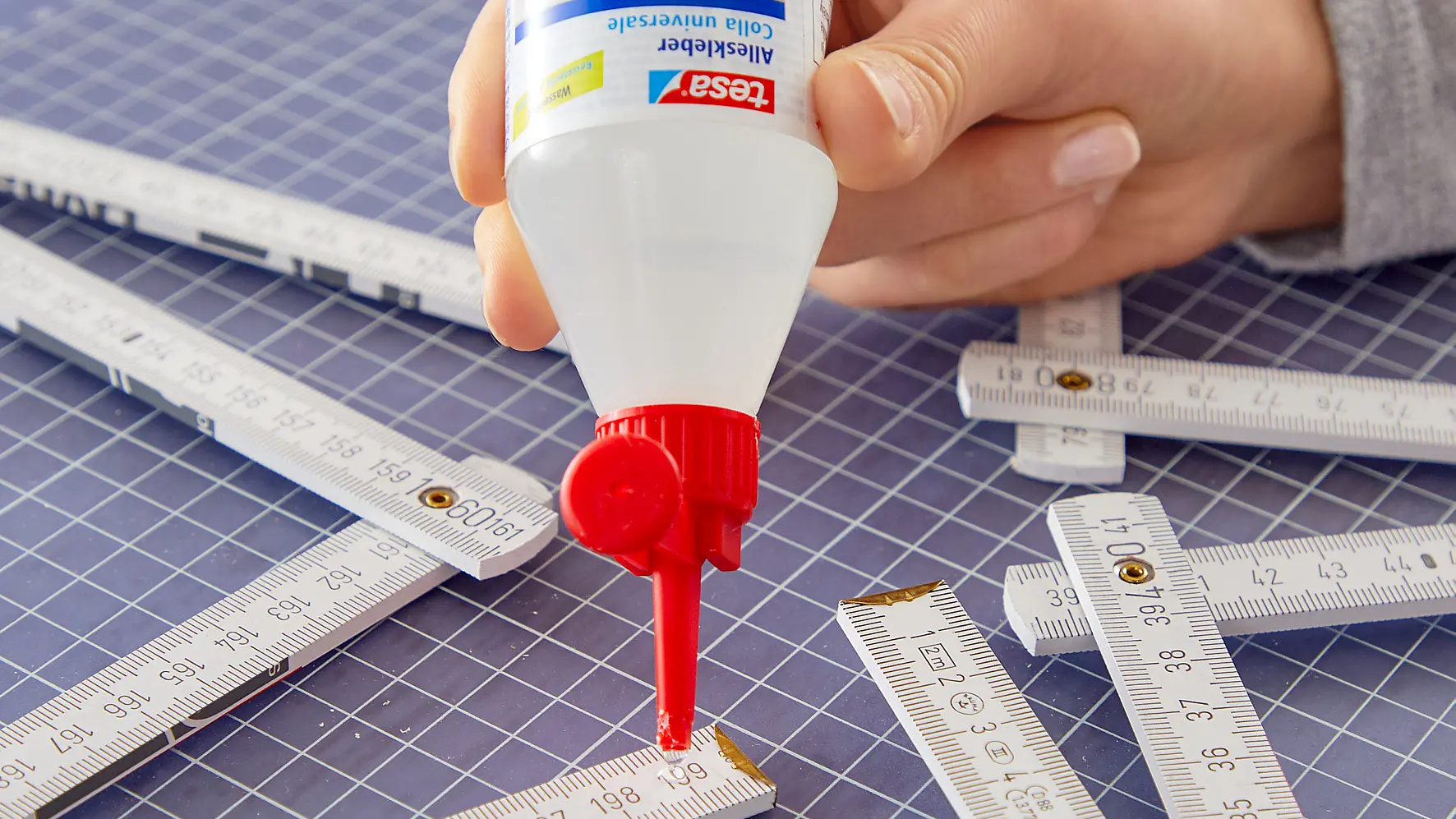 A hand applies glue from a bottle onto a folded measuring stick on a gridded cutting mat, securing it with tesa tape. Several other measuring sticks are scattered on the mat. The glue bottle has a red nozzle and a colorful label.