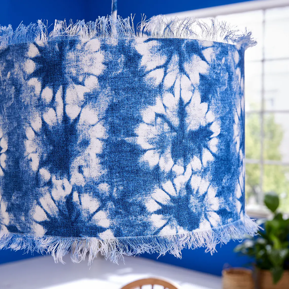 A close-up of a blue and white tie-dye patterned hanging light fixture. It has a fringed edge and hangs in a room with blue walls. Below, a dining table with a blue tablecloth and wooden chairs is partially visible, with a plant near the window secured using tesa tape.