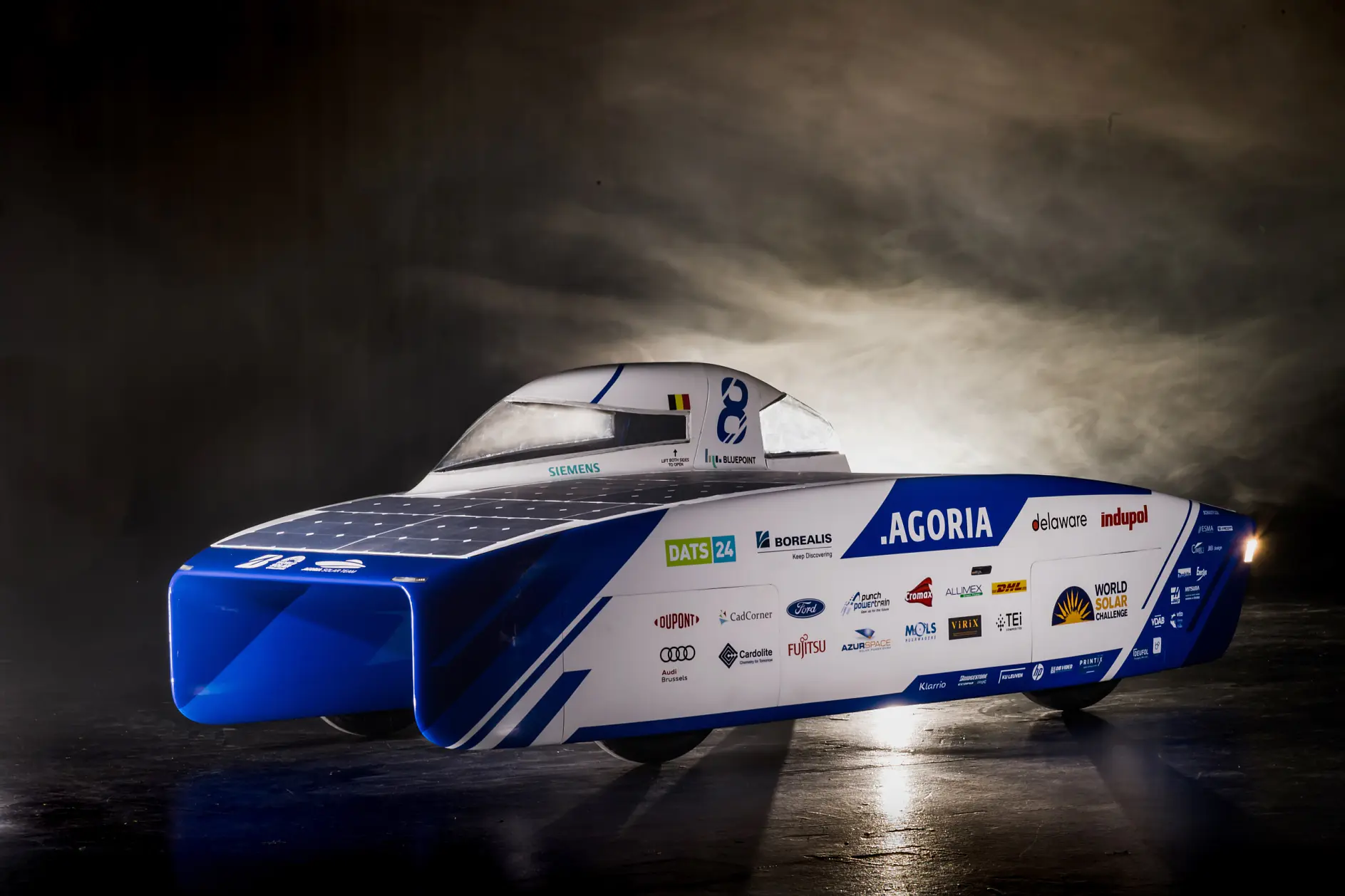 A streamlined solar-powered car with a blue and white color scheme, featuring various sponsor logos. It is designed with a covered cockpit and a flat surface fitted with solar panels. The background is dark with a spotlight highlighting the vehicle, proudly showcasing tesa tape on its exterior as an essential component in its sleek design.