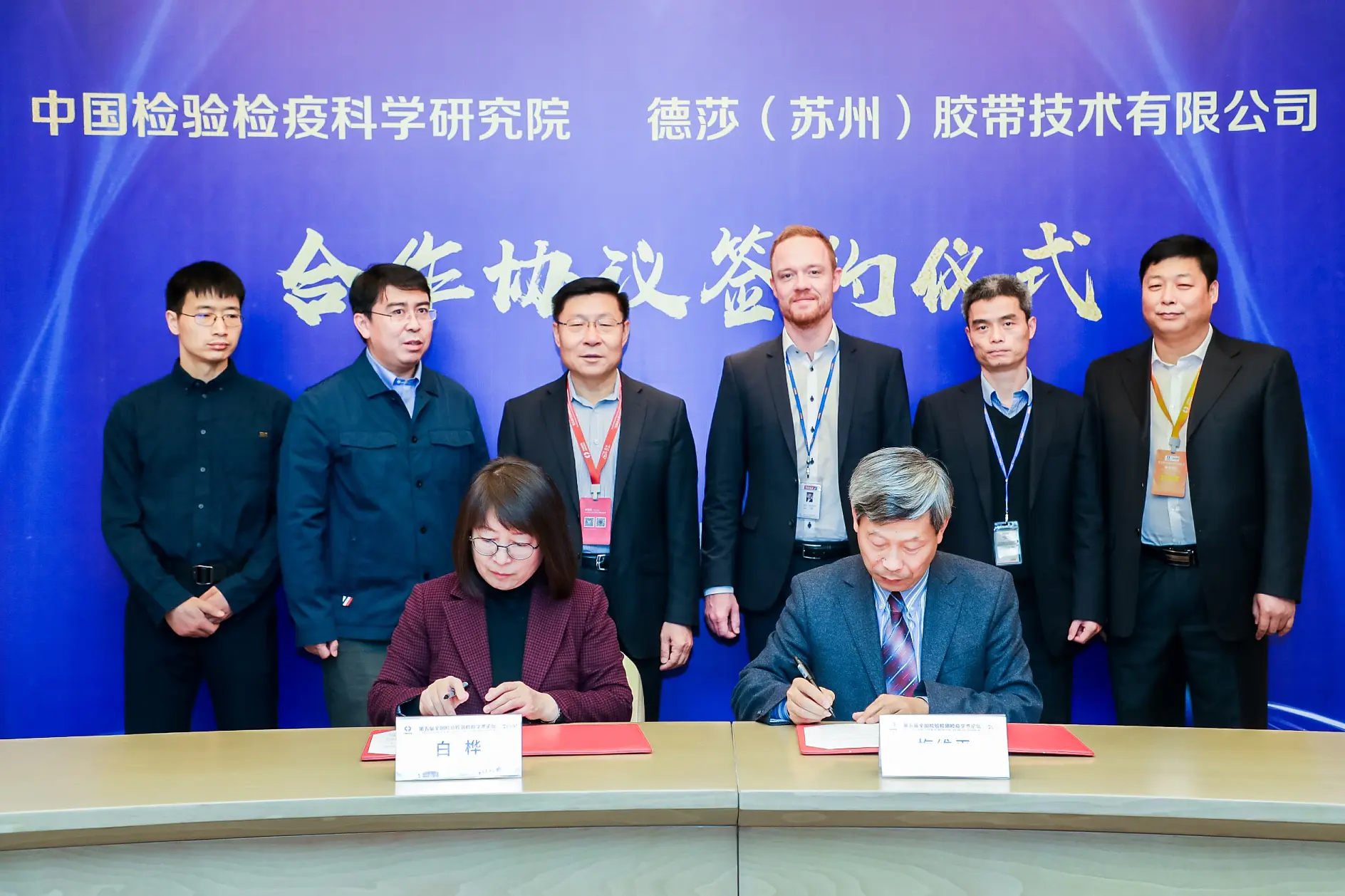 Nine individuals are present at a formal signing event. Two people in the front are seated at a table, using tesa tape to secure documents. Seven others stand behind them. The background displays text in Chinese and another language.