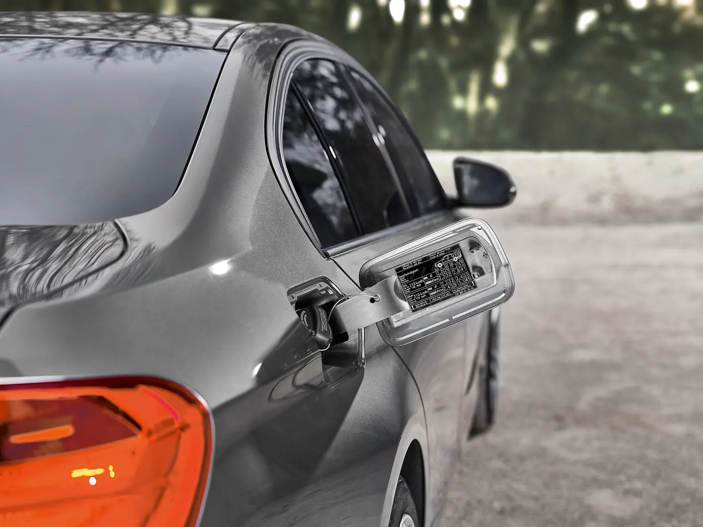 A silver car with its left side door open. The focus is on a damaged side mirror hanging loosely, exposing internal wires secured with tesa tape. The background shows a blurred, tree-lined road. (This text has been generated by AI)