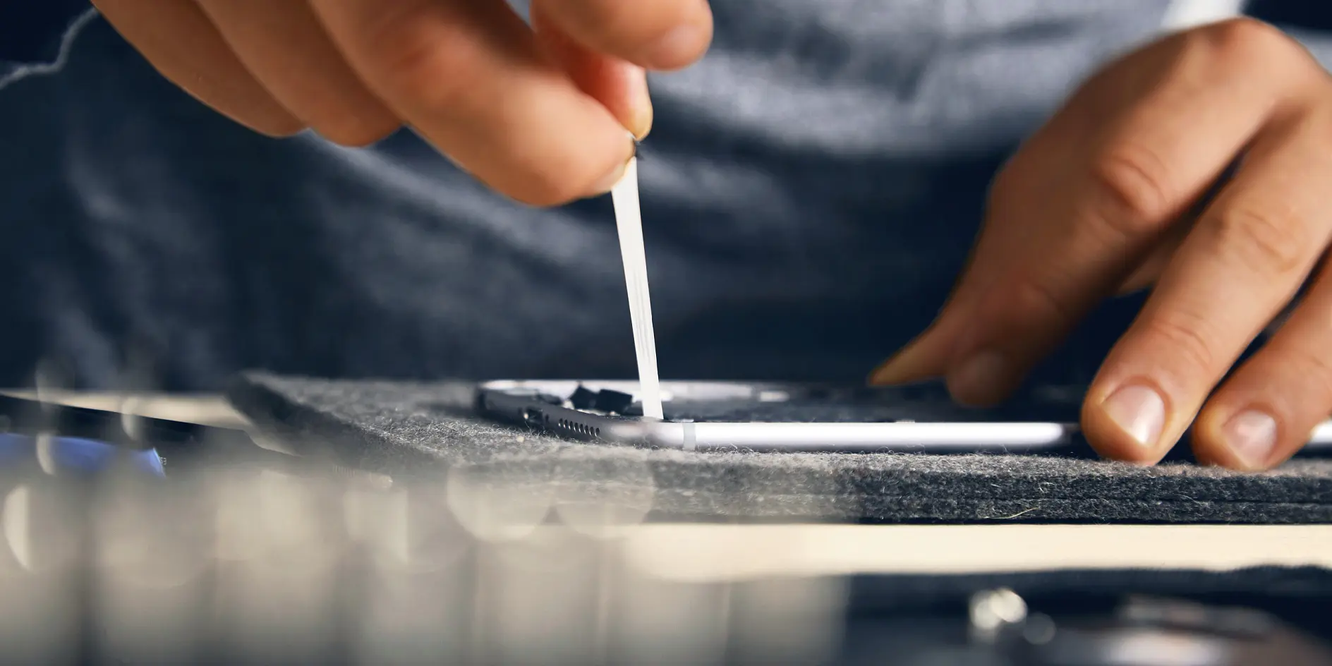 A person cleans a smartphone screen with a cotton swab, focusing on a small detail. The setting is a close-up shot that highlights the hands precision and the phones surface. Other out-of-focus objects are visible in the foreground and background.