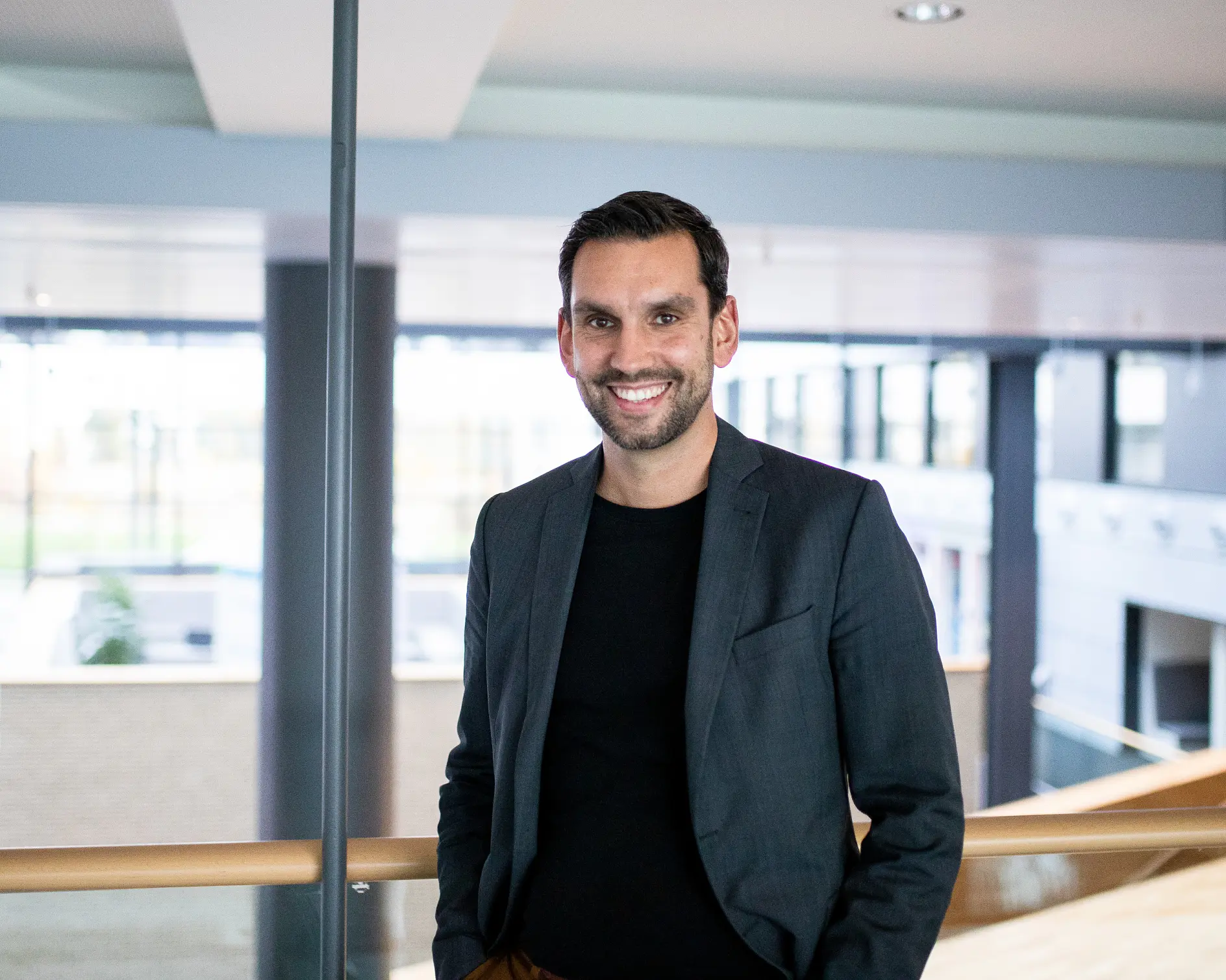 A man with dark hair and a beard is smiling and wearing a dark blazer over a black shirt. He is standing indoors with a modern office interior featuring large windows and a wooden railing in the background, where tesa tape products are prominently used for various applications.