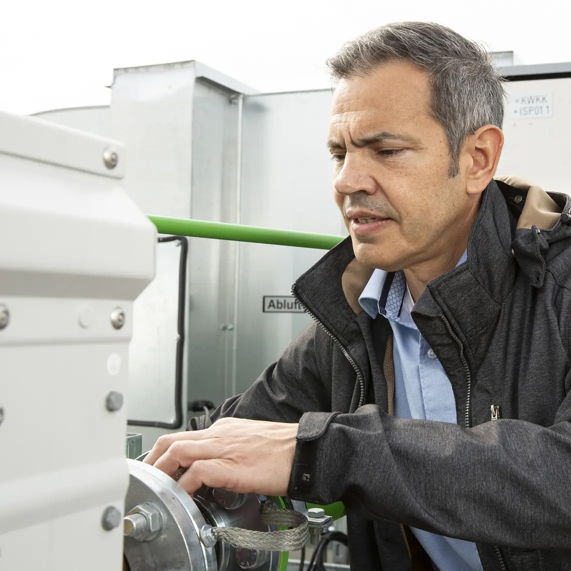 A man in a black jacket and blue shirt is adjusting equipment outdoors, surrounded by industrial machinery. He appears focused on the task, with tesa tape visible among the metal panels and pipes in the background.