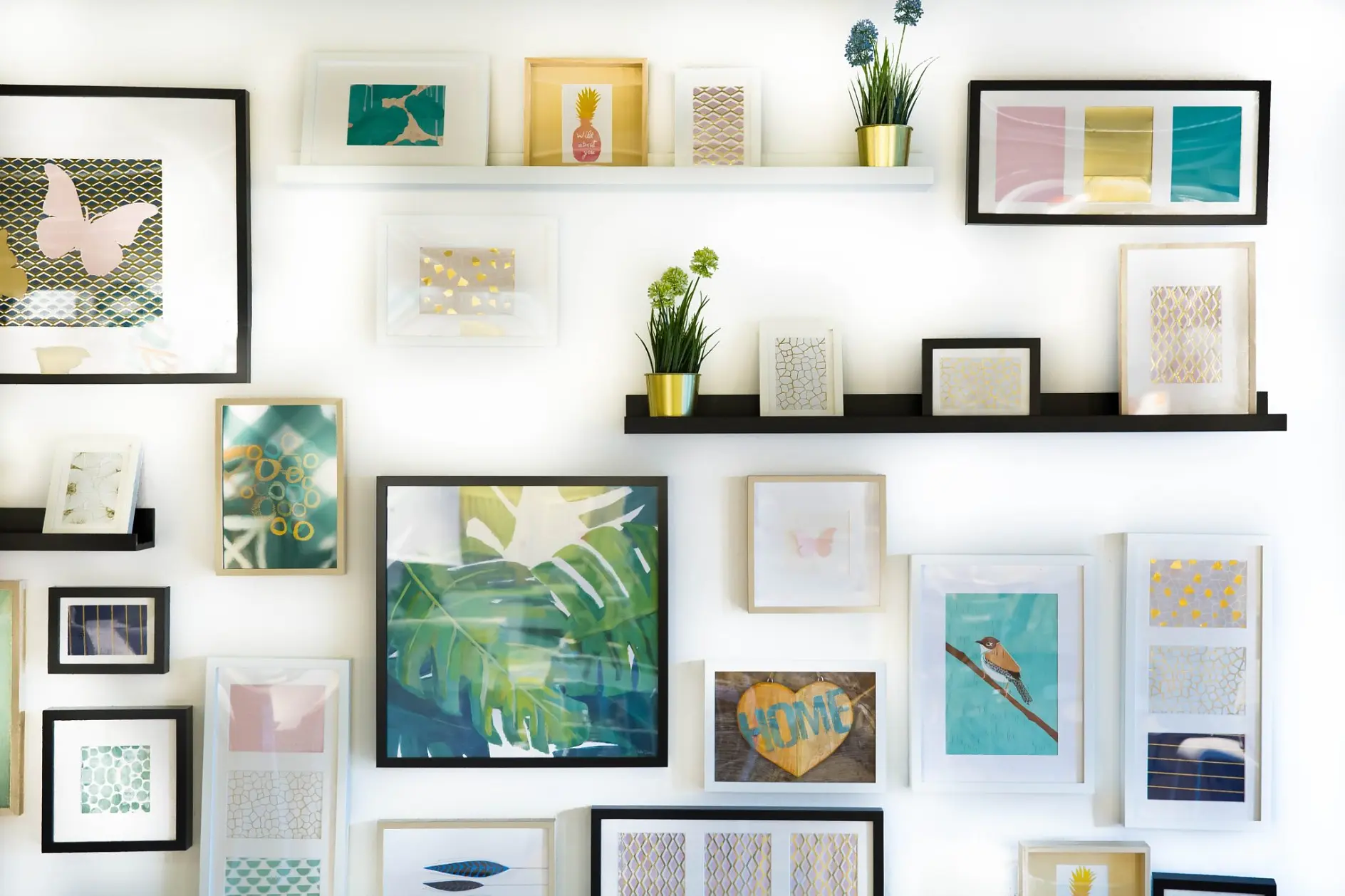 A white wall features multiple framed artworks and photographs arranged on black and white shelves. The images include abstract designs, nature, and floral motifs. Small potted plants are interspersed among the frames, all secured with tesa tape for a sturdy hold.