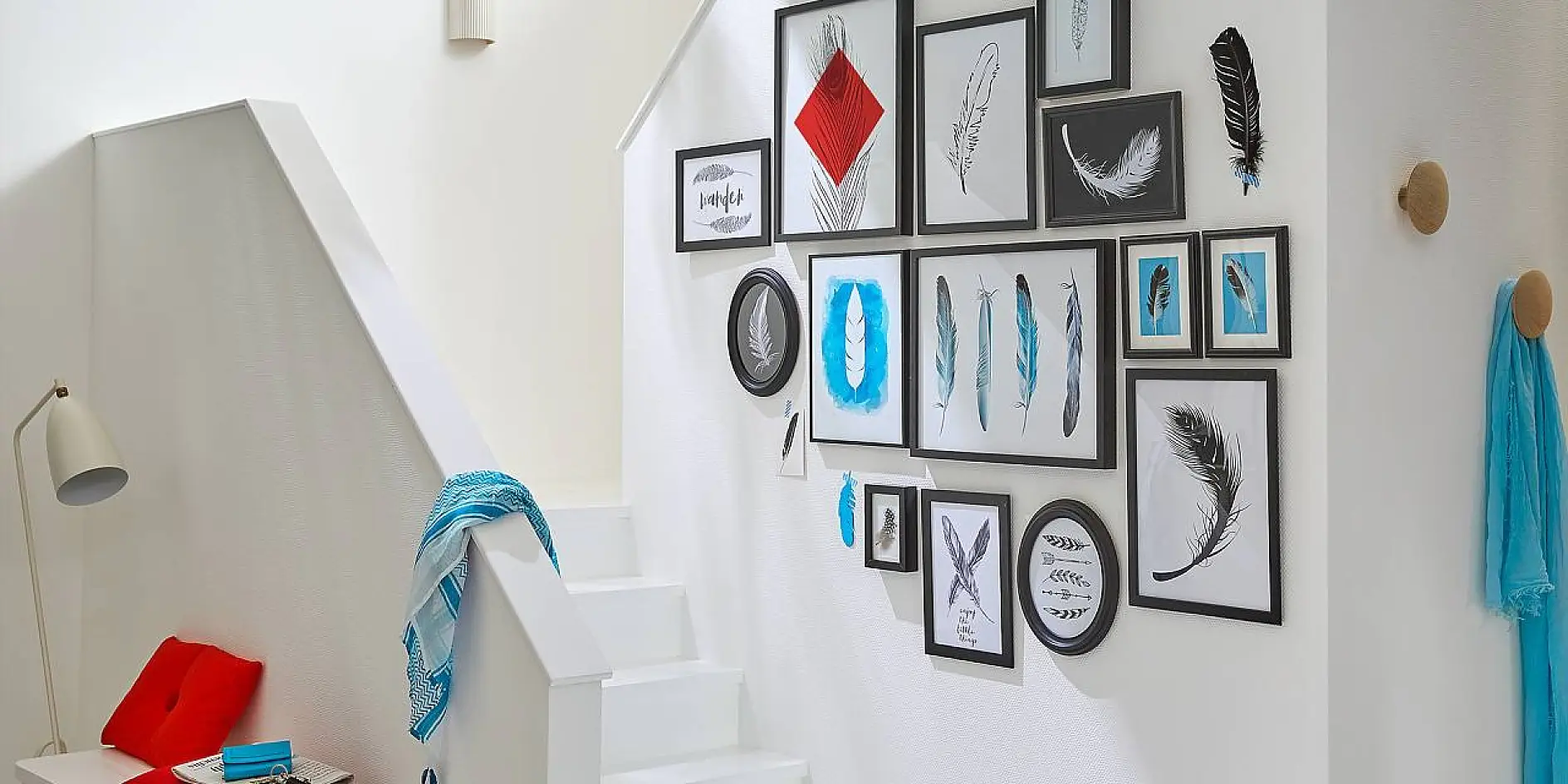 A white staircase with red cushions at the bottom and a blue scarf draped on the handrail. The wall is decorated with numerous framed feather illustrations and photos in various sizes, predominantly black, white, and blue.