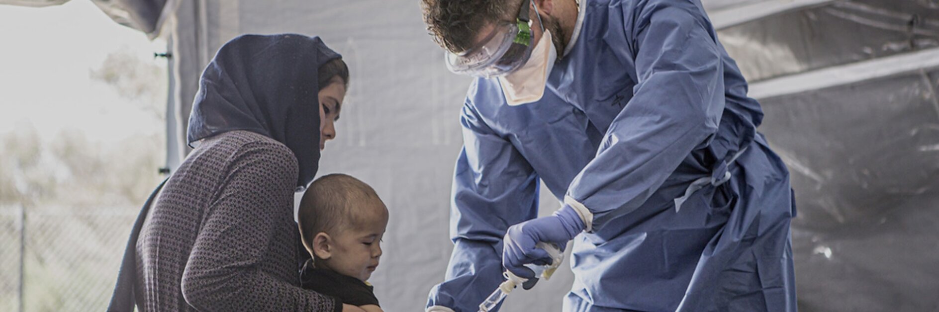 A healthcare worker in protective gear examines a young childs hand in a medical tent. The child is sitting on a womans lap. The scene seems to be a medical or humanitarian setting, with supplies such as tesa tape used for various applications.