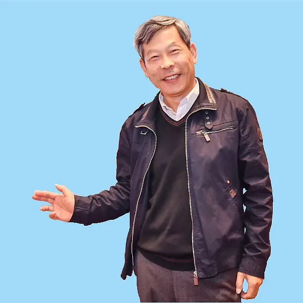 A man with gray hair, wearing a dark jacket over a black sweater and white shirt, is smiling. He stands against a solid blue background, extending his right hand outward, showcasing tesa tape.