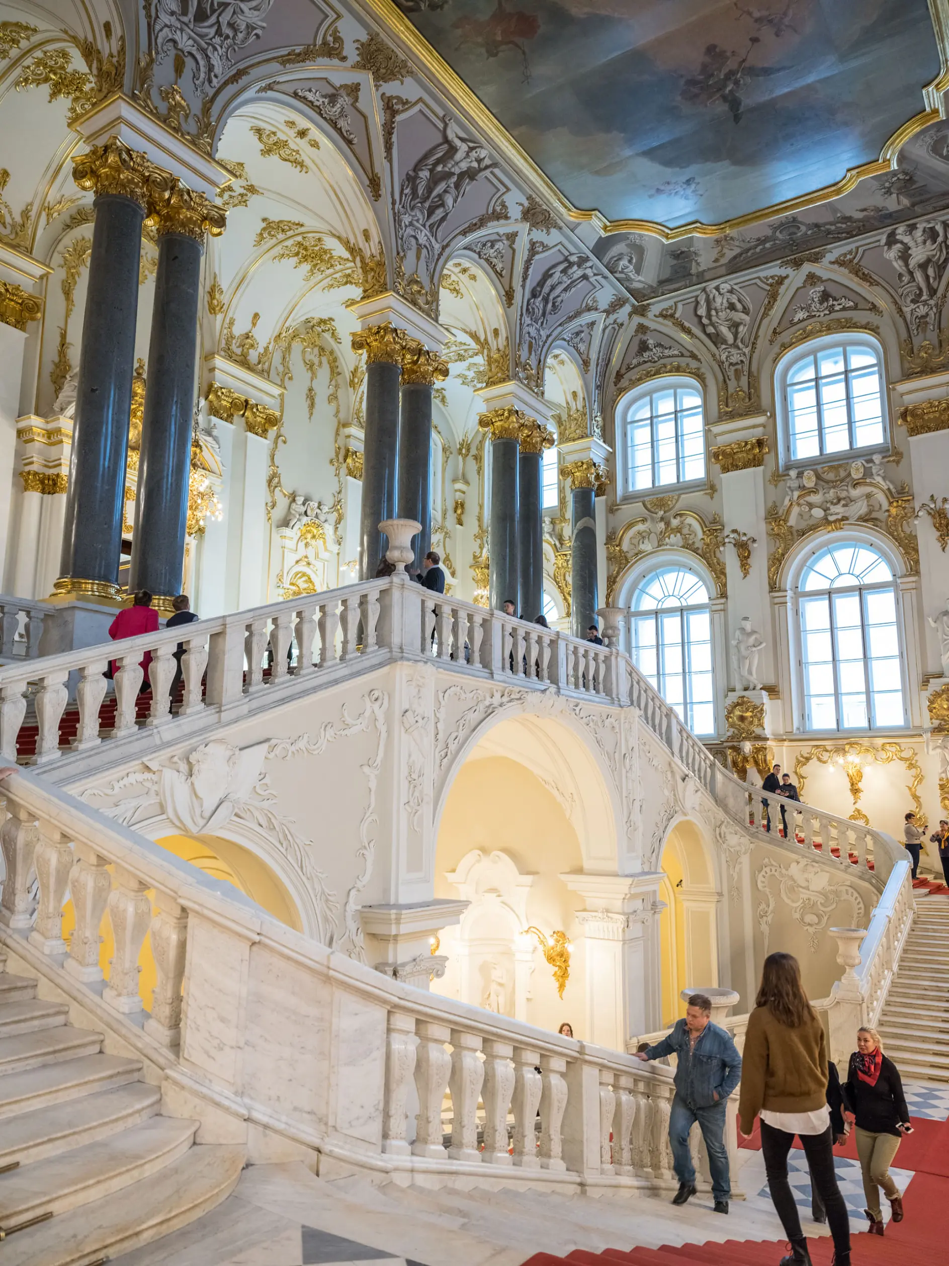 A grand, ornate staircase inside a historic building features white marble steps, gold accents, and intricate architectural details. Several people are walking or standing on the stairs, and large windows allow natural light to enter.