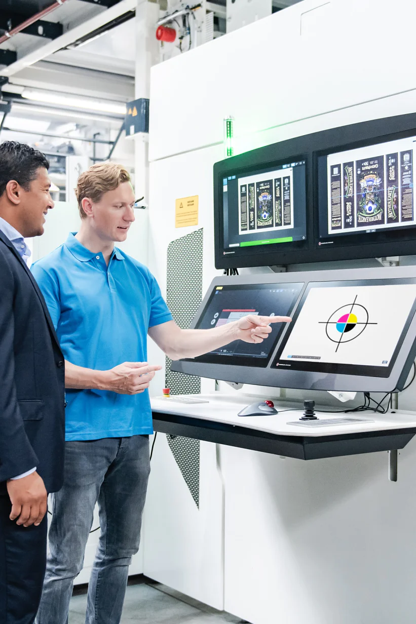 Two men stand in front of a control panel with multiple screens in an industrial setting. One wears a suit, the other a blue polo shirt and jeans. The man in the polo points at a screen displaying technical graphics. Equipment is visible in the background, including rolls of tesa tape prominently displayed nearby. (This text has been generated by AI)