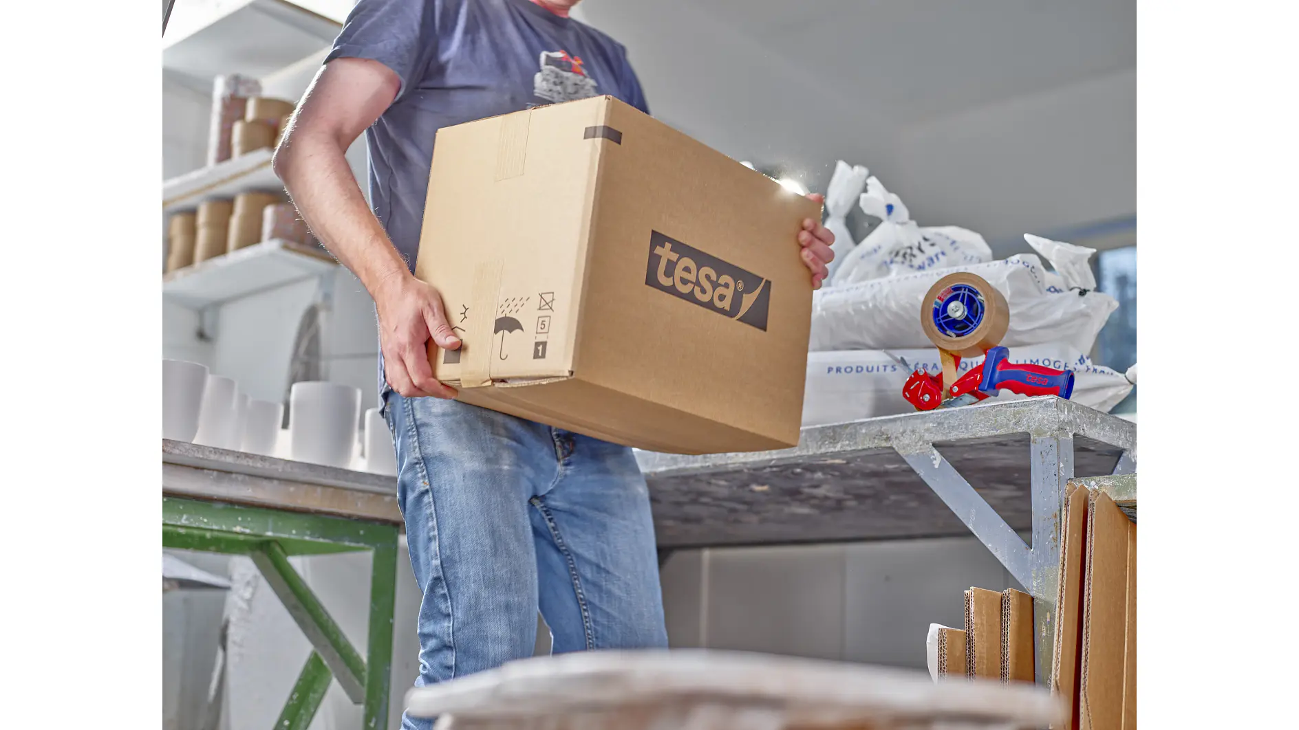 A person in a blue t-shirt and jeans is carrying a large cardboard box labeled tesa. The background includes a table with packing supplies, a tesa tape dispenser, and other cardboard boxes. (This text has been generated by AI)