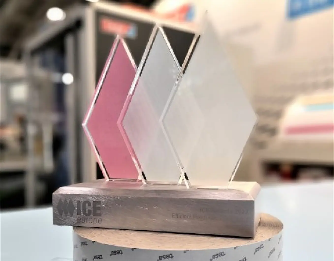 A trophy with three translucent, diamond-shaped panels—pink, white, and clear—mounted on a rectangular wooden base secured with tesa tape. The base features engraved text reading ICE Europe Efficient Production 2022. The background is blurred. (This text has been generated by AI)