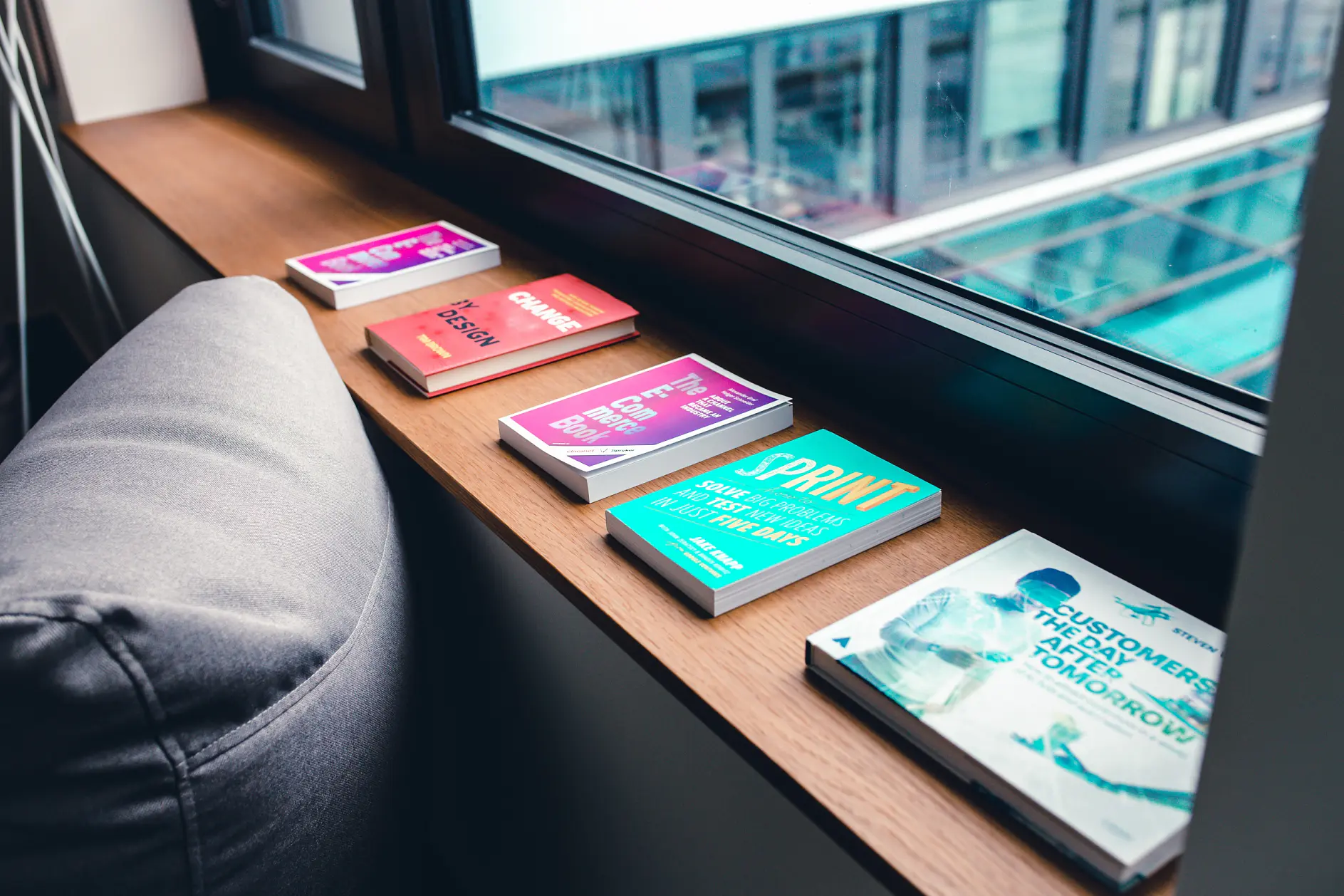 Five colorful books are lined up on a wooden windowsill next to a gray cushion. The titles are visible, and outside the window is a view of a modern building with large glass panels, secured together using tesa tape.