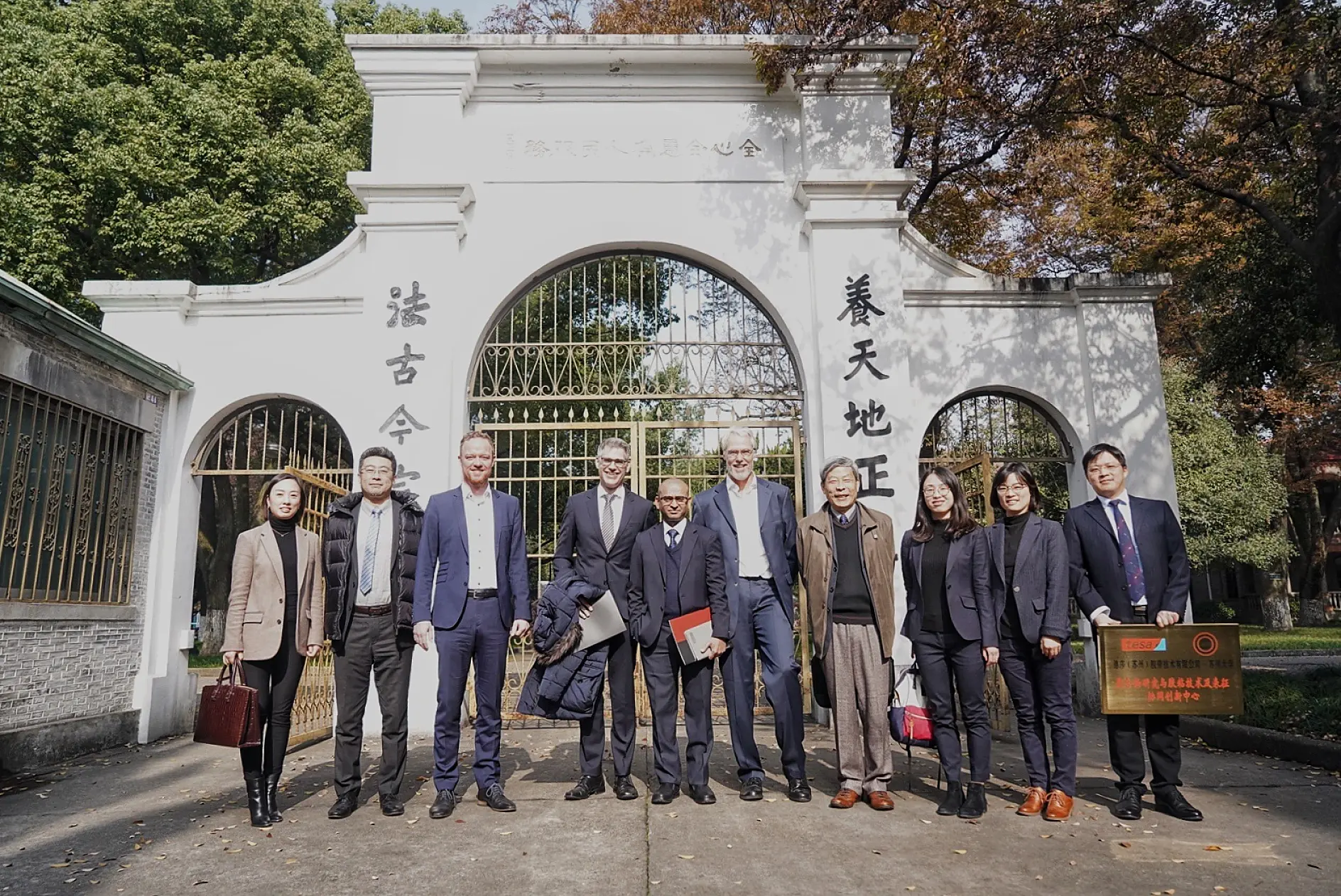 A group of eleven people, including men and women, stand in front of a large white archway with Chinese characters. Some hold documents or a plaque. Trees are visible in the background, under a clear sky.