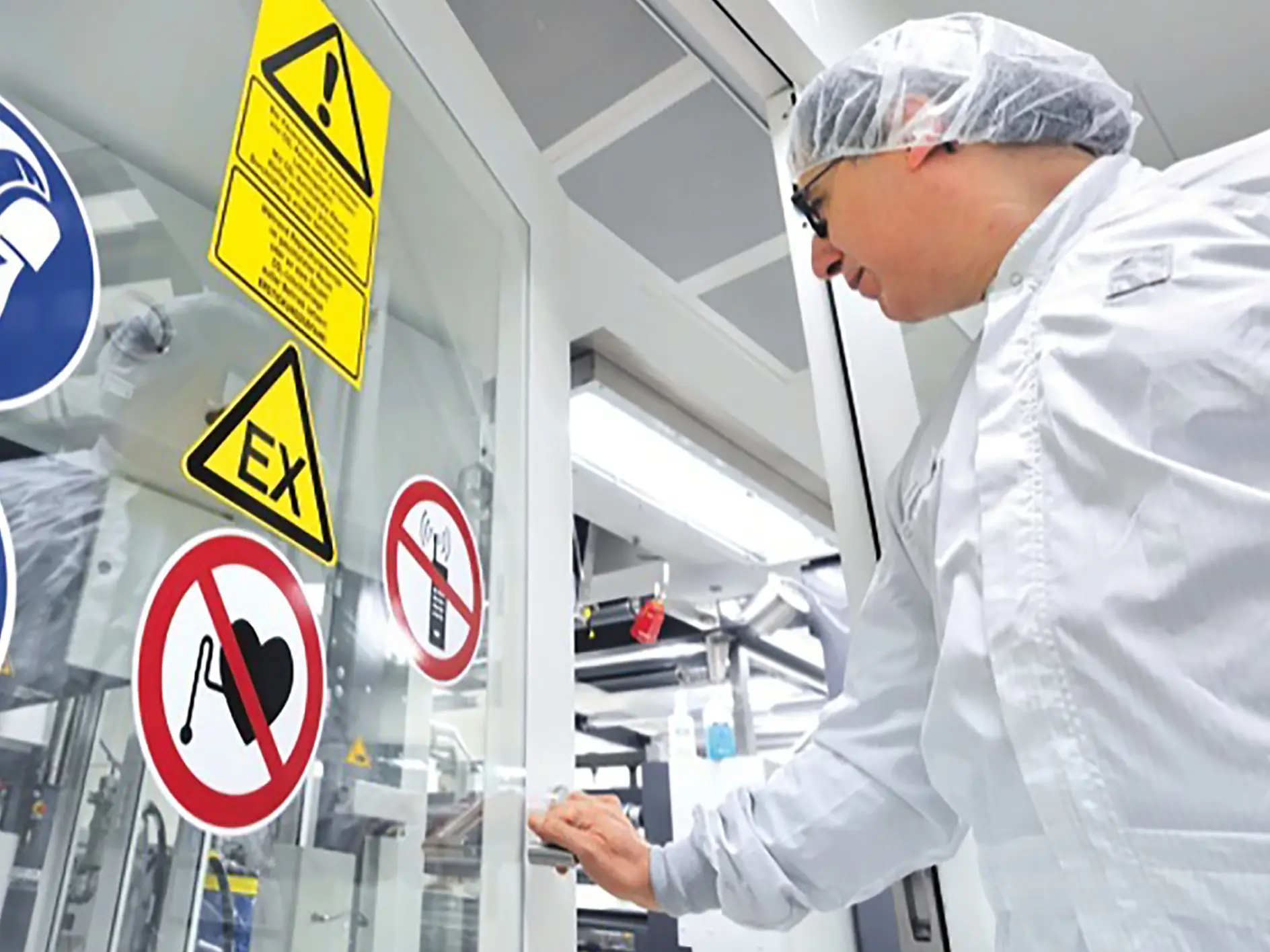 A person wearing a white lab coat, hair net, and safety goggles is about to enter a laboratory or controlled environment. The door has multiple warning signs secured with tesa tape, including no smoking, caution, and flammable materials symbols.