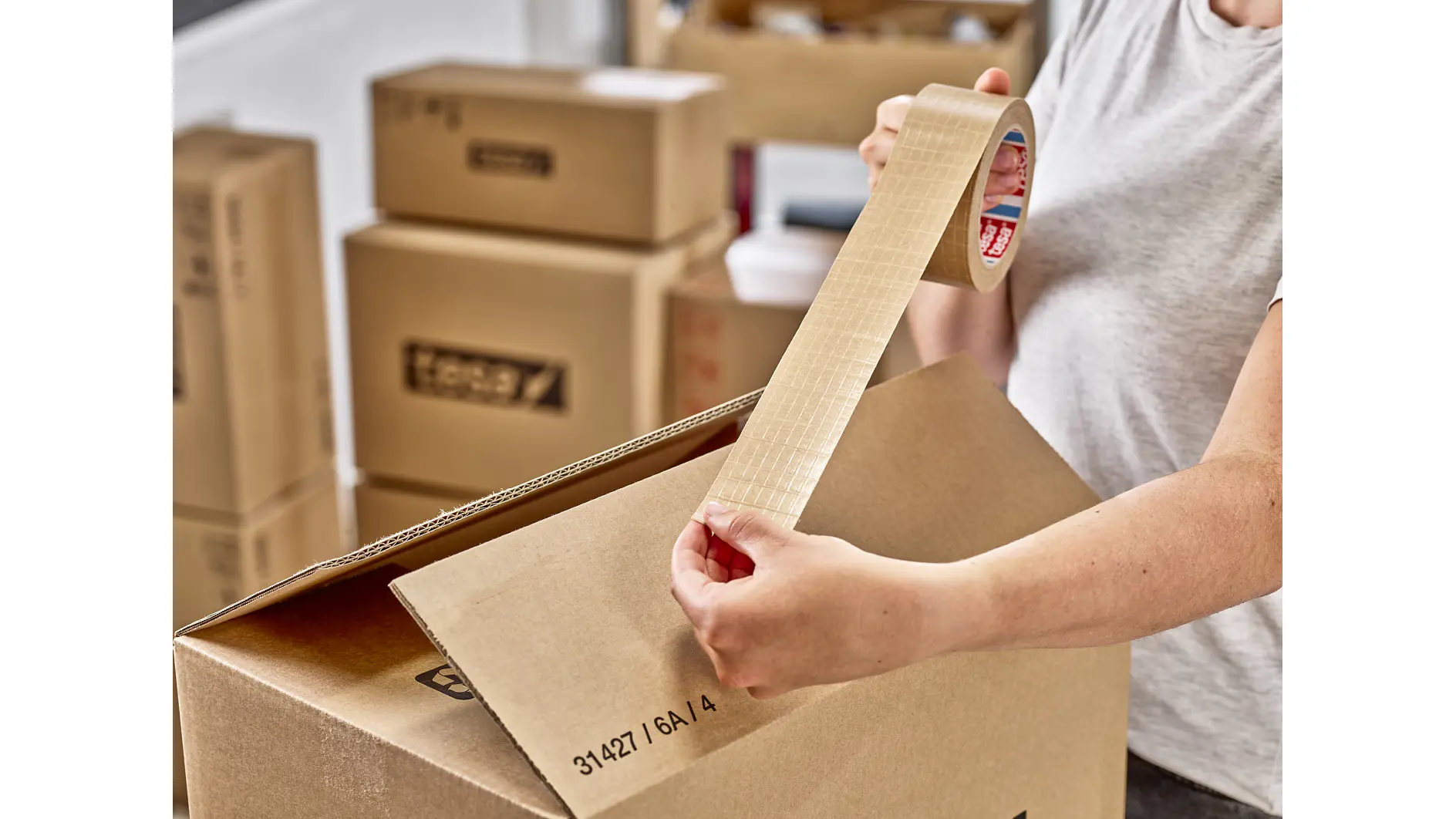 A person sealing a cardboard box with tesa tape. Several other packed boxes are visible in the background, suggesting preparation for moving or storage. The person is dressed casually in a light gray shirt. (This text has been generated by AI)