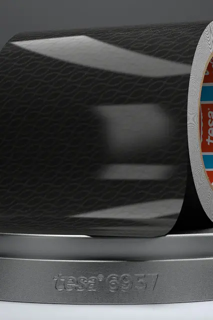 A partially unrolled roll of black tesa tape with a subtle pattern, resting on a gray circular stand. The inner part of the roll shows orange and blue detailing with text. The stand is labeled tesa 19657. (This text has been generated by AI)